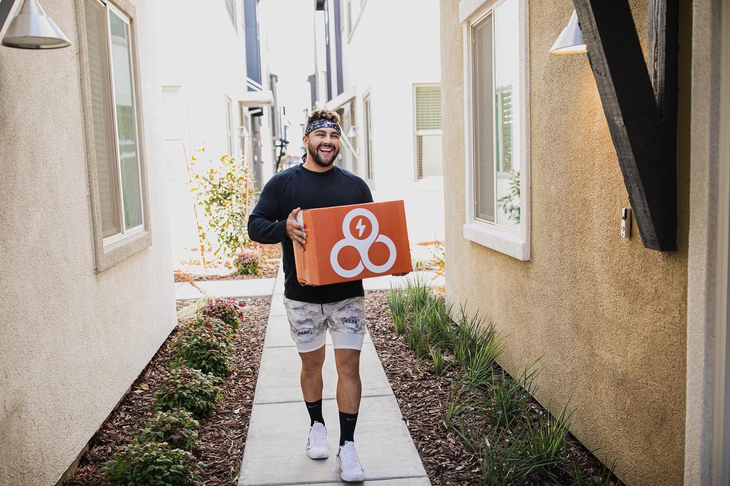 A man smiling and holding an orange box with a white Trifecta logo, walking on a sidewalk between two beige stucco houses.