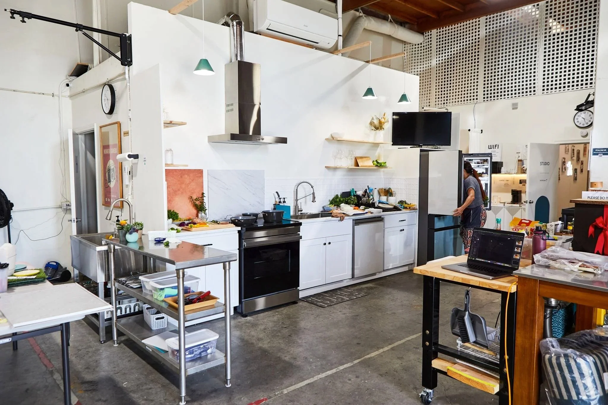 A modern industrial-style kitchen with white cabinets, a stove, and open wooden shelves, with a woman working near the refrigerator.