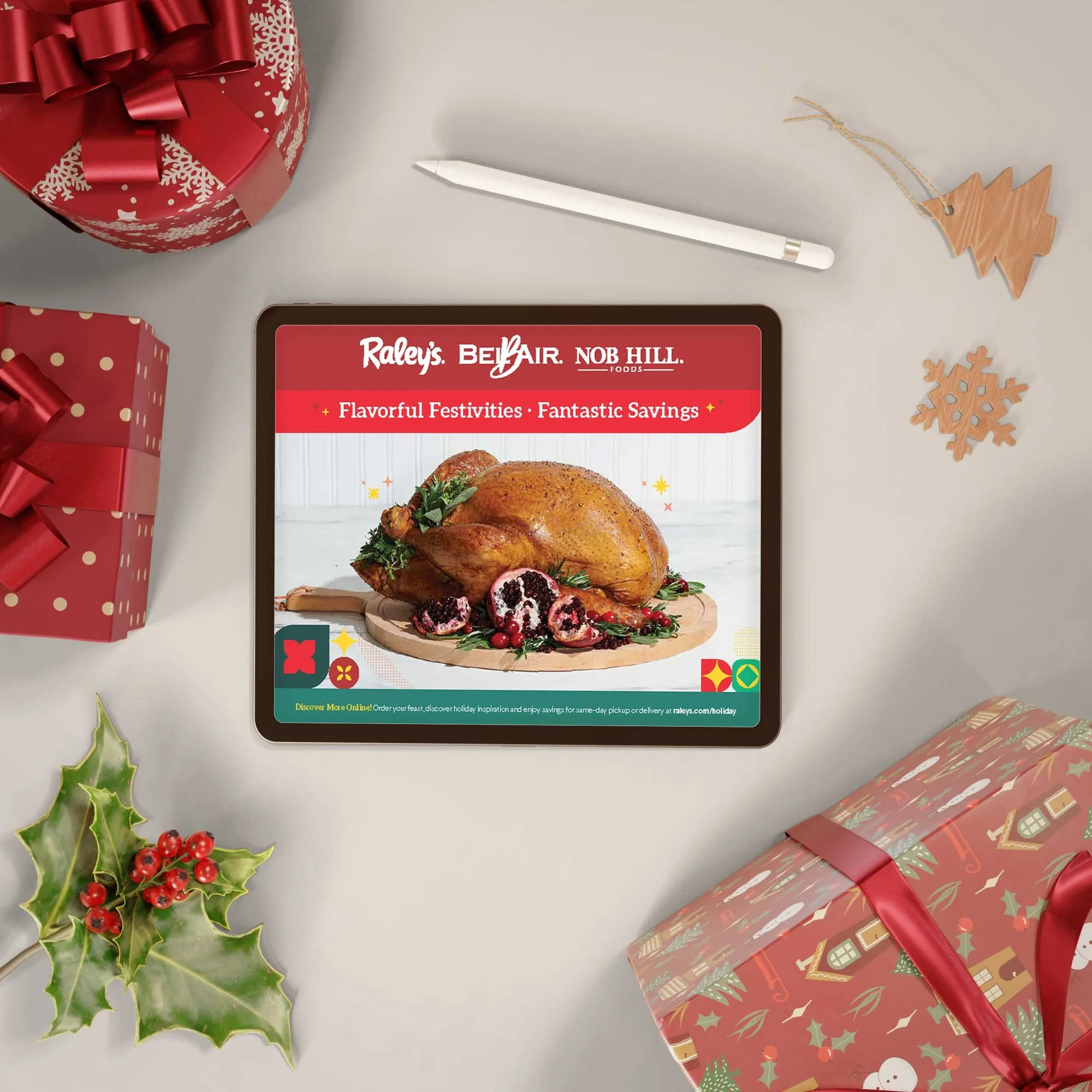 A digital tablet displaying an advertisement for holiday foods, with a roasted turkey on a wooden cutting board and a festive background. The scene includes Christmas presents, holly, snowflake and Christmas tree decorations, and a white pen on a light-colored surface.