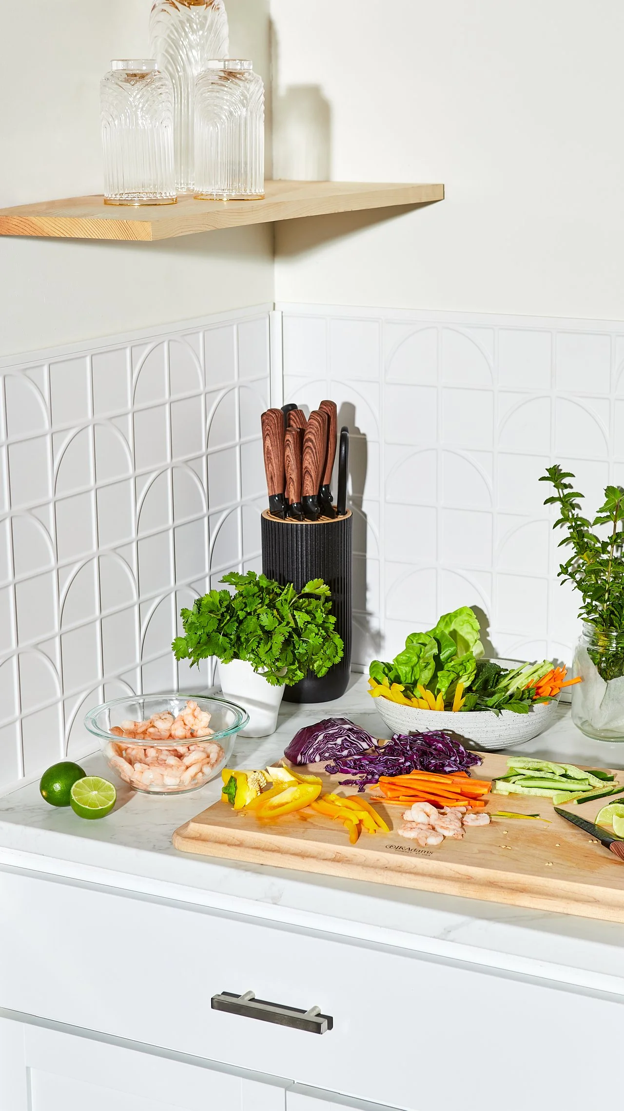 Kitchen countertop with vegetables, a bowl of raw shrimp, a lime, and kitchen knives in a holder.