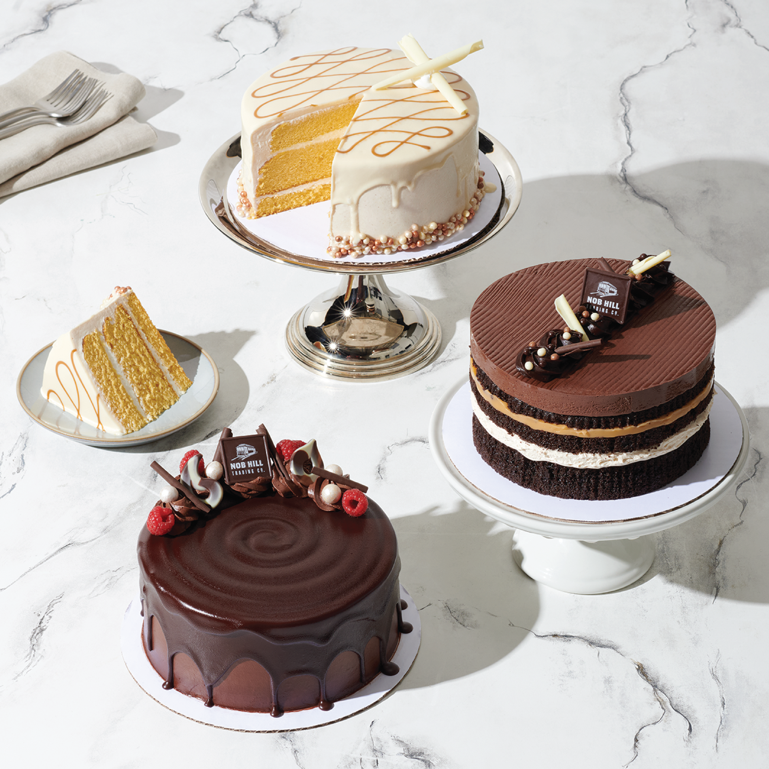 Assorted decorated cakes on a marble surface, including chocolate and vanilla layers, with toppings like chocolate pieces, raspberries, and white chocolate curls.