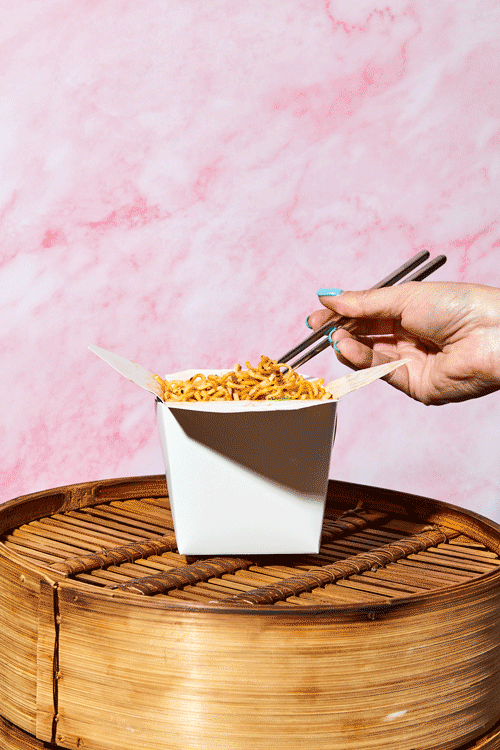 Hand using metal chopsticks to pick up noodles from a white takeout container, resting on a round bamboo tray, with a pink textured background.