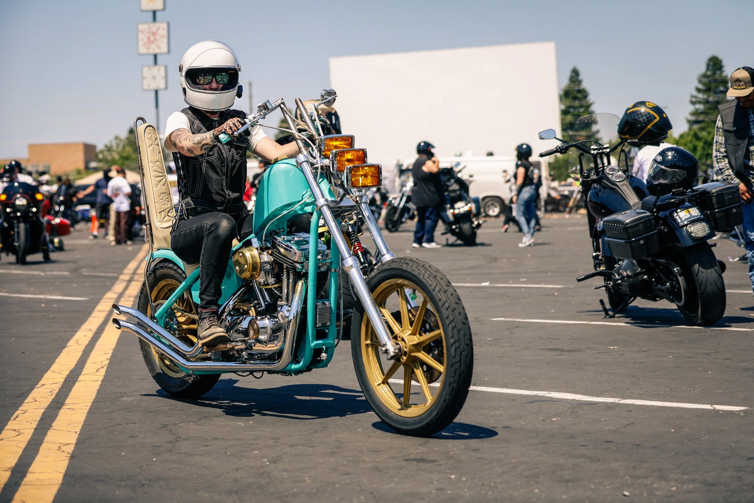 Person wearing a helmet riding a teal and gold motorcycle in a parking lot with other motorcyclists in the background.