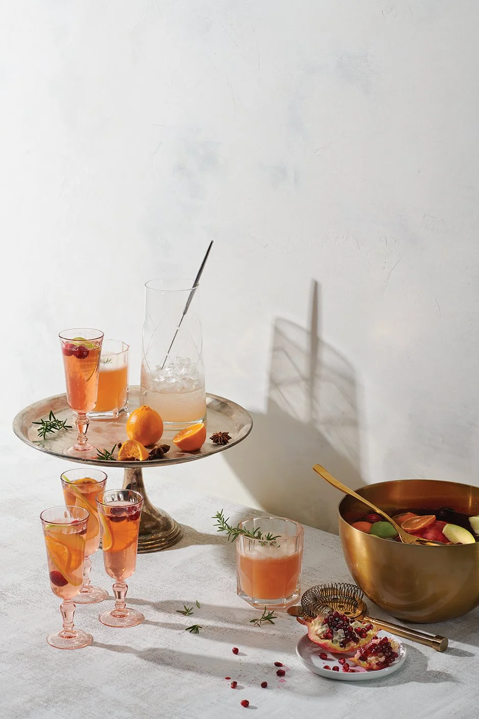 A table with assorted drinks, including pink cocktails with fruit garnishes, a glass with ice and a straw, and a large bowl filled with sliced fruit, with some pomegranate seeds and sprigs of herbs scattered on the table.