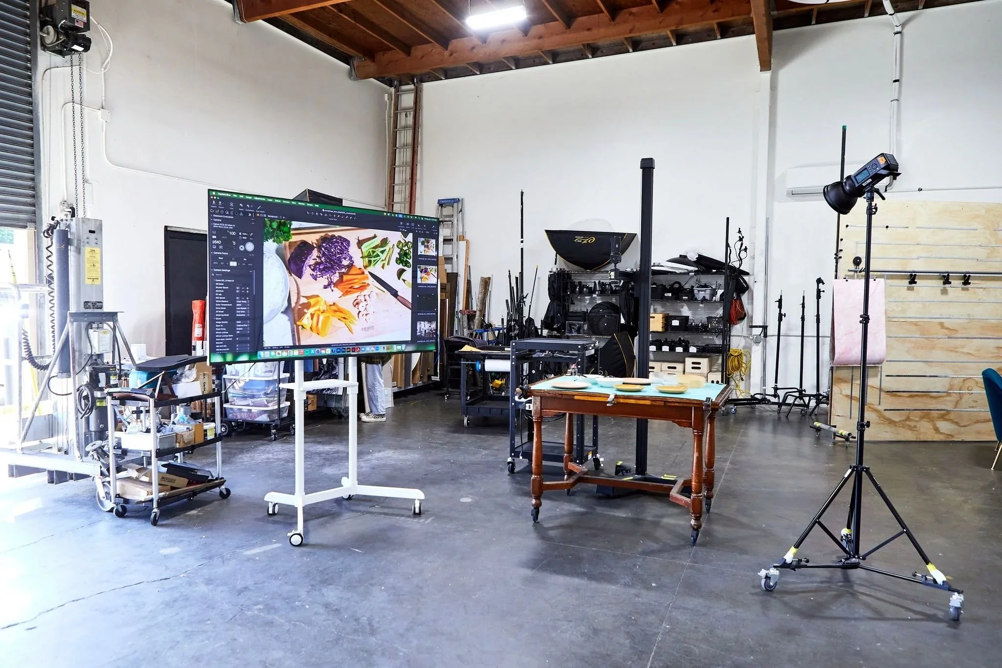 Photography studio with a large monitor displaying food photography, various lighting equipment, a wooden table, and shelves with camera gear in a spacious room with a concrete floor and wooden ceiling beams.