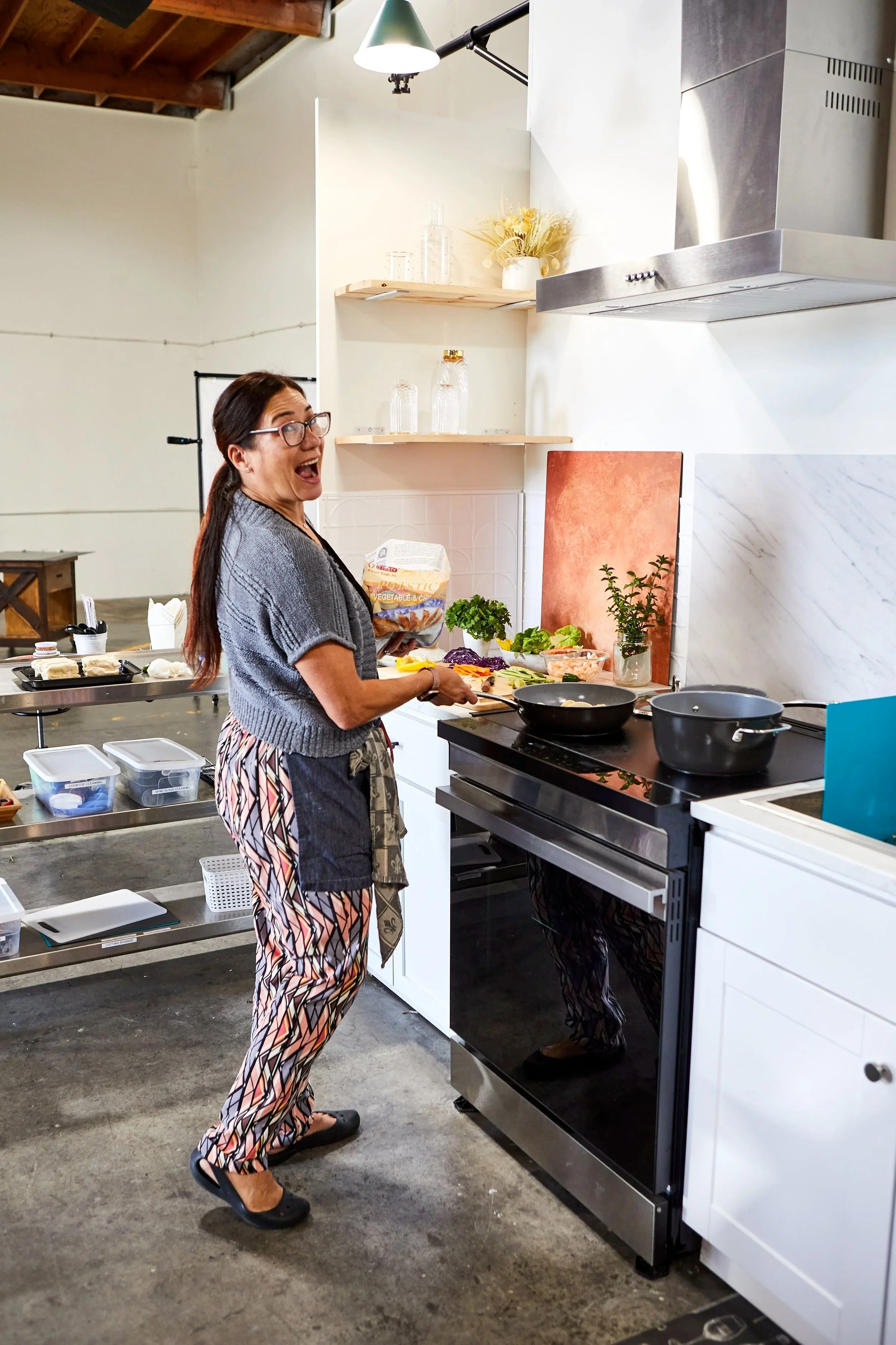 Woman in a kitchen cooking and smiling while holding a bag of vegetables or chips.