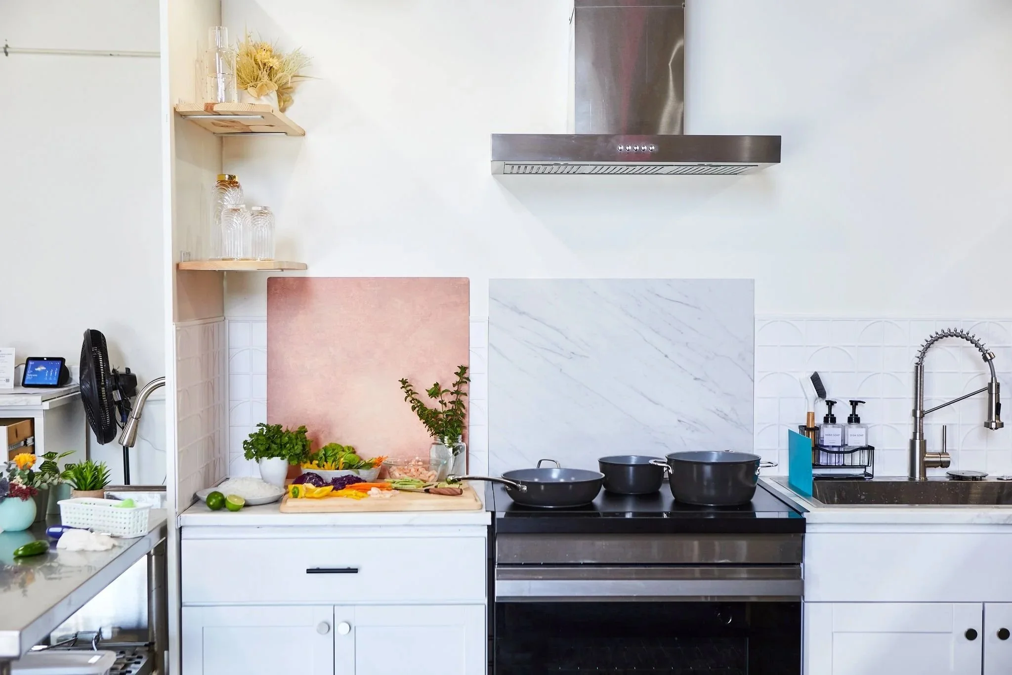 A modern kitchen with a white countertop, black stove with three black pots, a stainless steel range hood, a sink with a high-arch faucet, and wooden shelves with glassware and decorative items. Vegetables and herbs are visible on the counter and a pink and white backsplash.