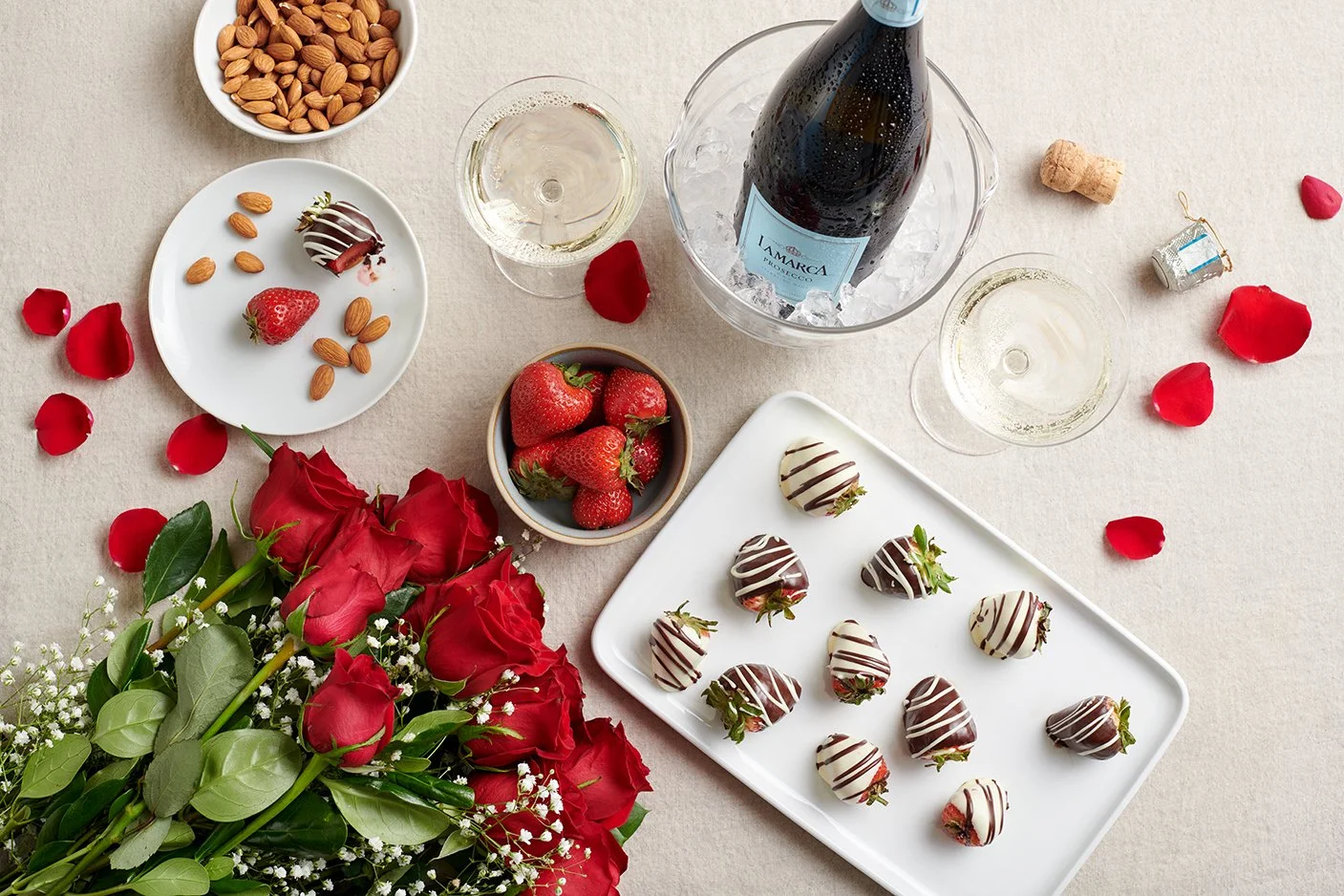 A table setting with chocolate-covered strawberries, a bowl of strawberries, almonds, two glasses of champagne, a bottle of wine in an ice bucket, rose petals, and a bouquet of red roses with white filler flowers.