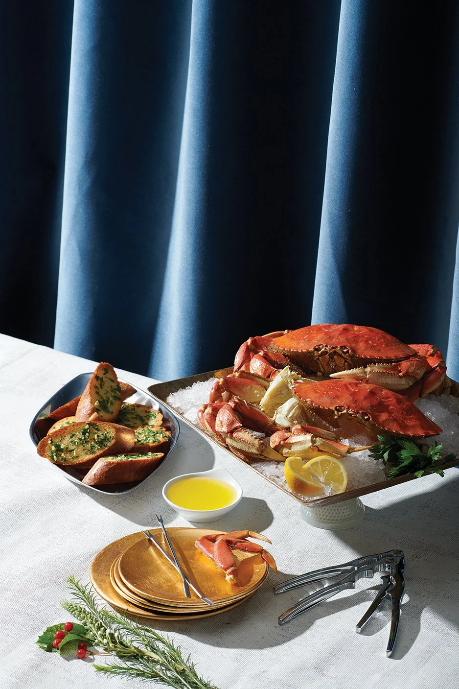 A seafood platter featuring crab on a bed of ice, garlic bread slices with herb butter, a lemon wedge, a small bowl of melted butter, and a sprig of parsley, with a blue curtain in the background.