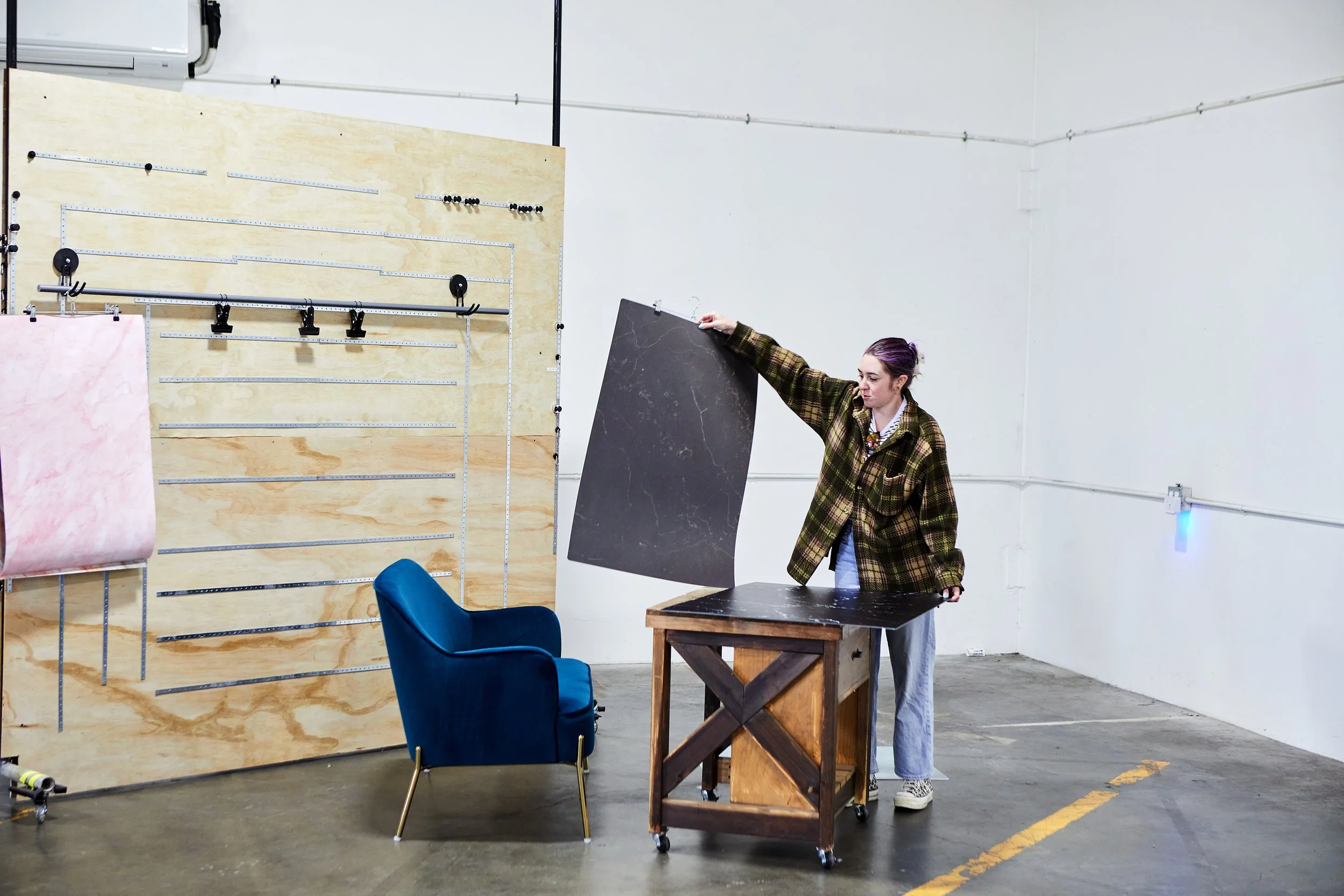 A woman is standing in an industrial-style room, holding a large black marble-patterned board, with a small wooden table and a blue armchair beside her. There is a large pegboard on the wall with various tools and materials hanging on it.