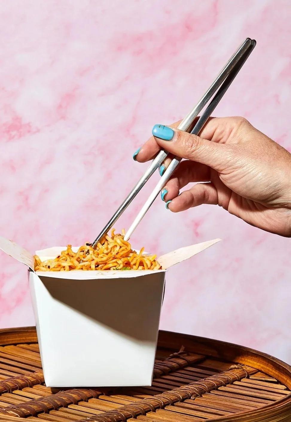 Hand holding metal chopsticks and a white plastic fork above a takeout box of noodles on a woven bamboo tray against a pink textured background.