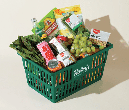 Green grocery basket filled with snacks and drinks, including grape bunches, leafy greens, canned drinks, bottled water, and snack packages, on a white surface.