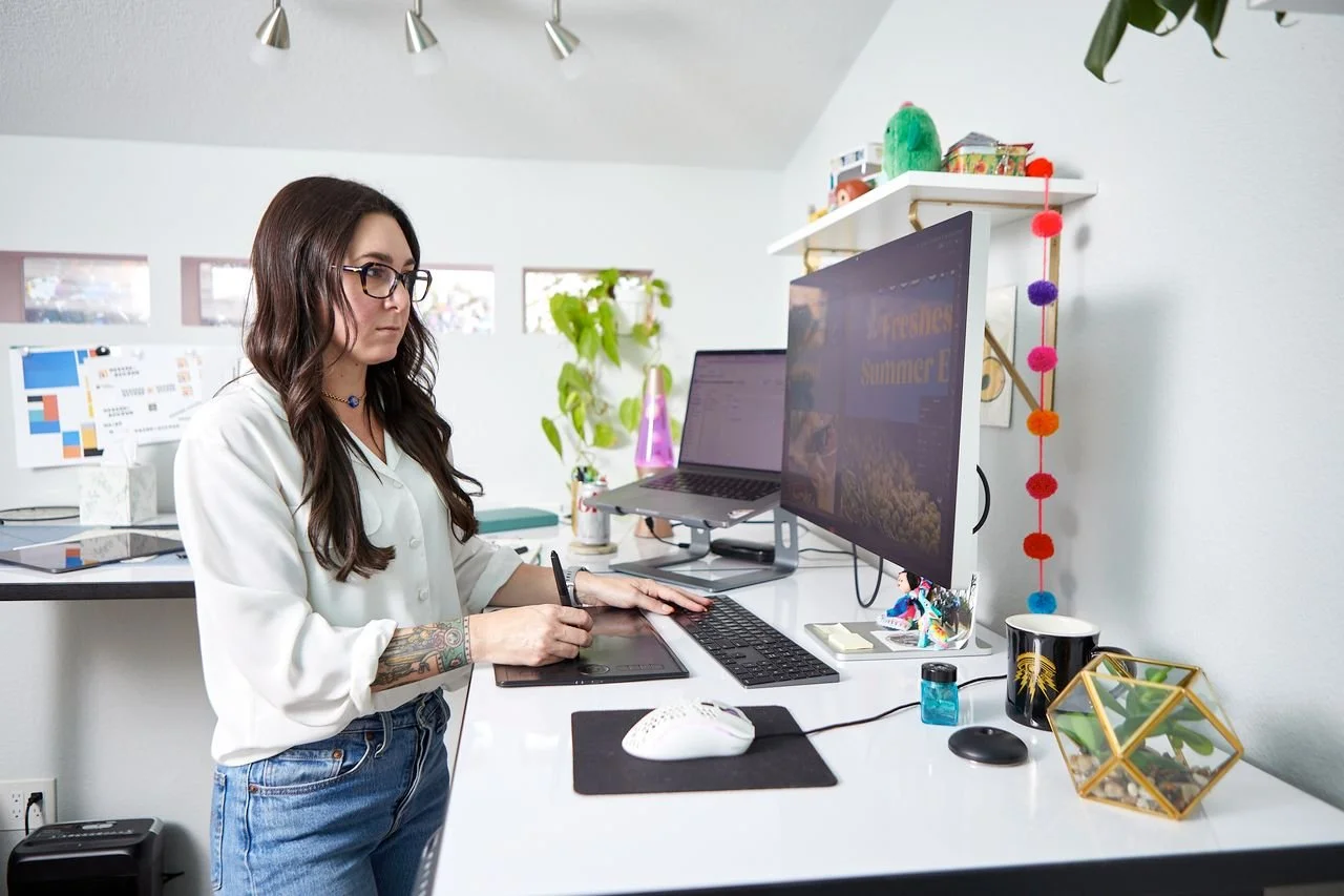 A woman with brown hair, glasses, and tattoos on her arm using a graphics tablet at a white desk, with a dual monitor setup, in a bright modern office with plants and colorful decor.