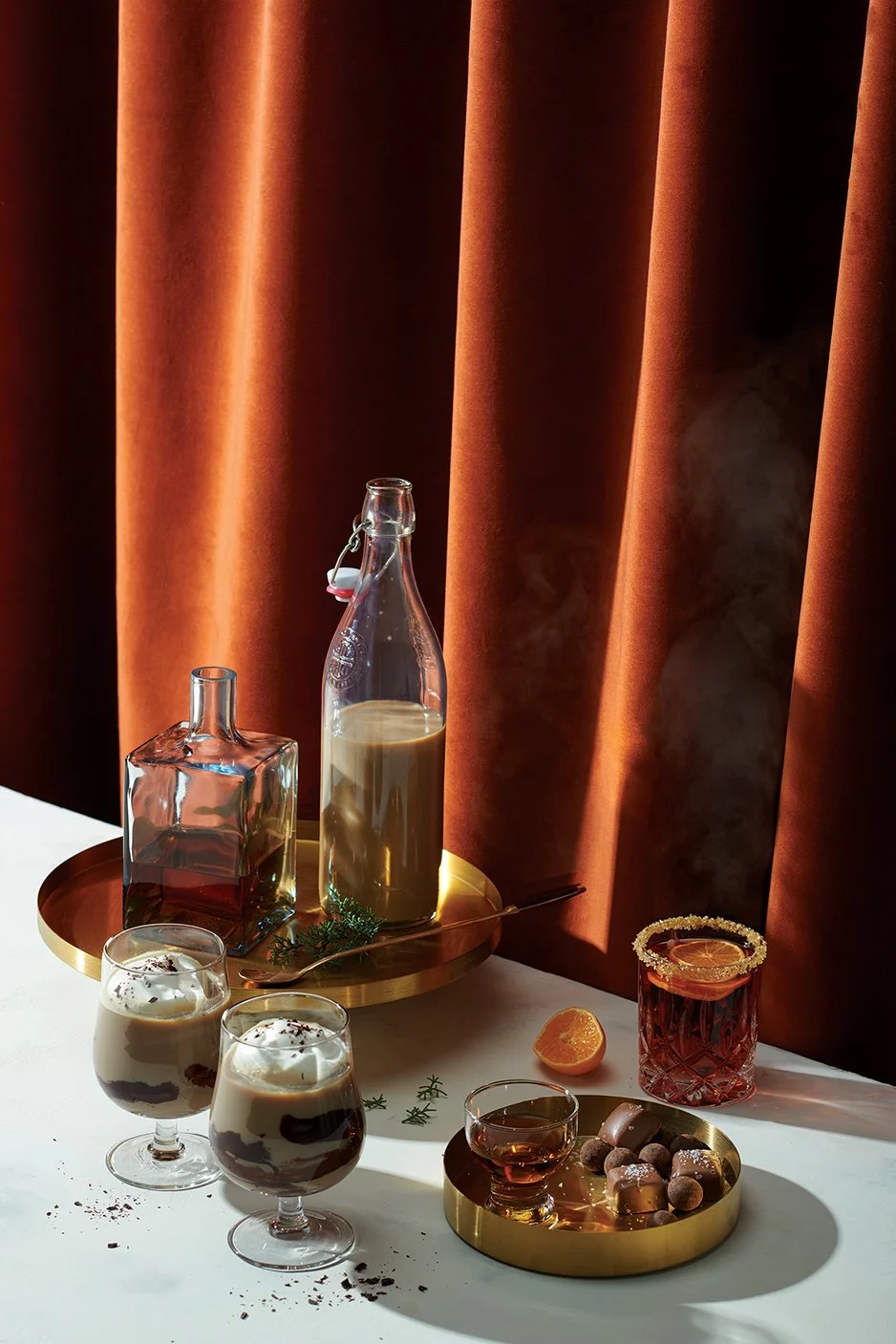 Decorative table setup with various drinks and desserts. There are two glasses of layered dessert in the foreground, a bottle of milk inside a glass bottle, an empty square glass bottle, a small cup of caramel, a glass of red-colored drink with a sal