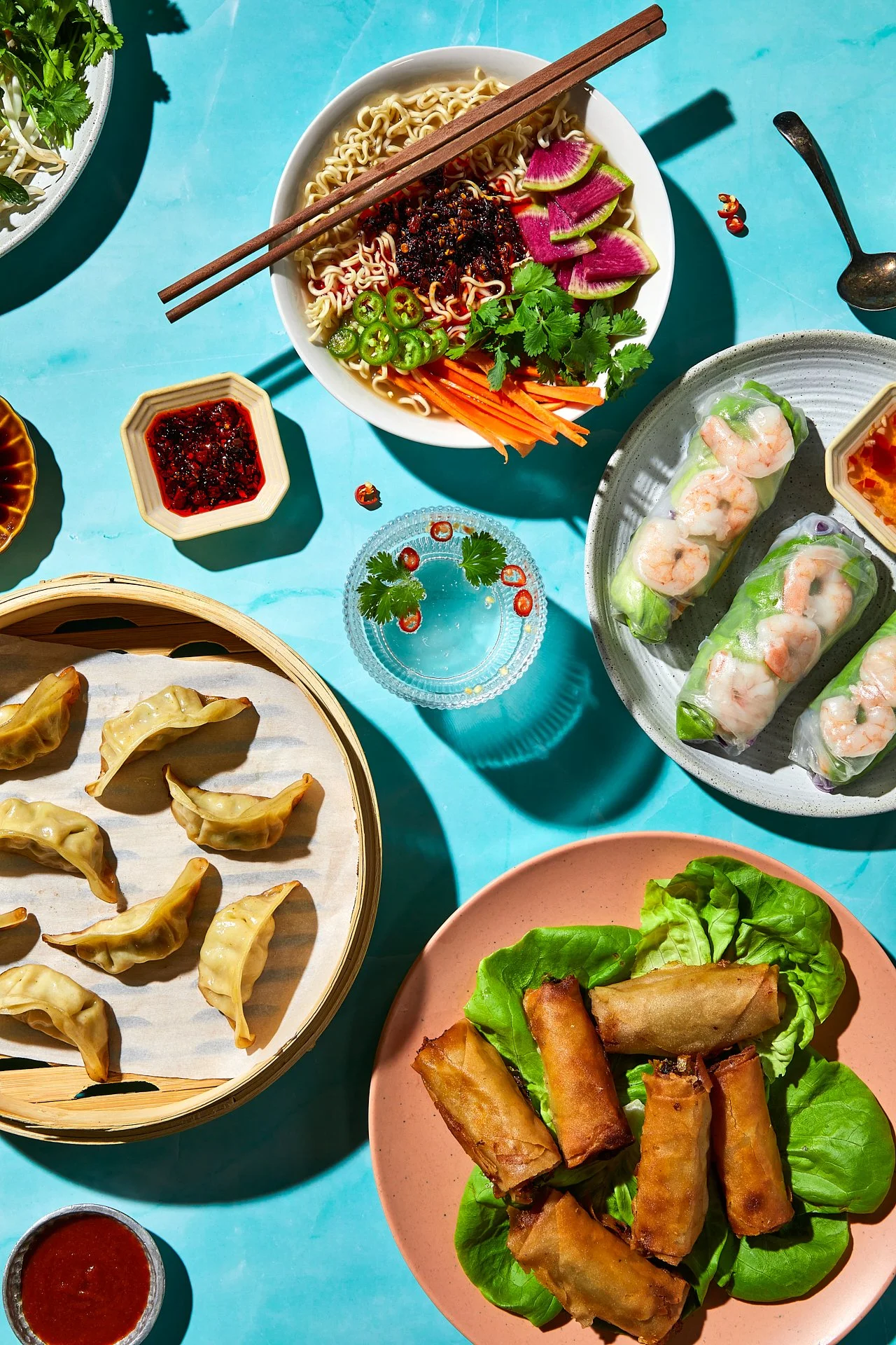 A table with Asian dishes including spring rolls on a pink plate, dumplings in a bamboo steamer, a bowl of ramen with vegetables and chopsticks, fresh vegetable spring rolls, dipping sauces, and a glass of water with chili and cilantro, all set on a 
