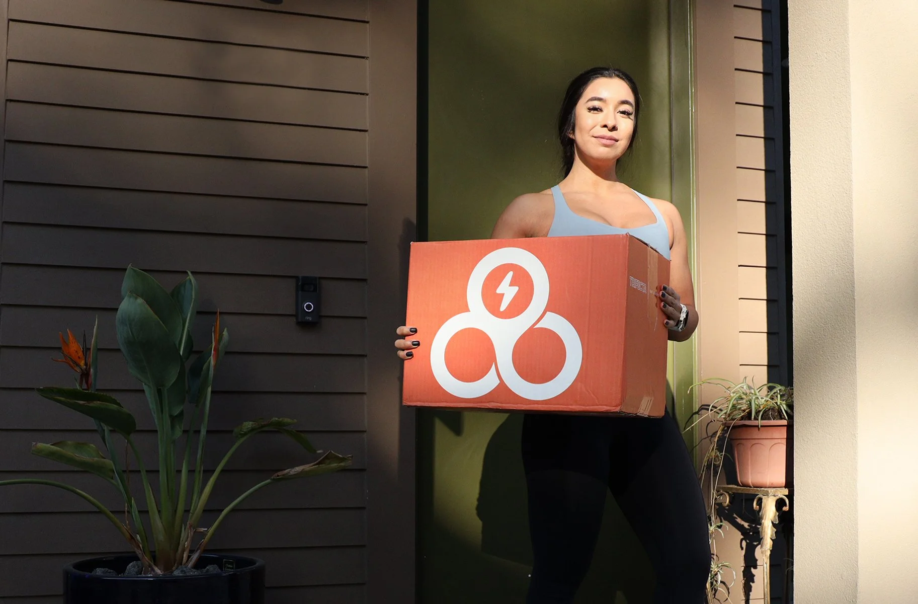 Woman standing at her doorstep holding a large orange box with a white logo, wearing a blue sports bra and black leggings, with potted plants nearby.