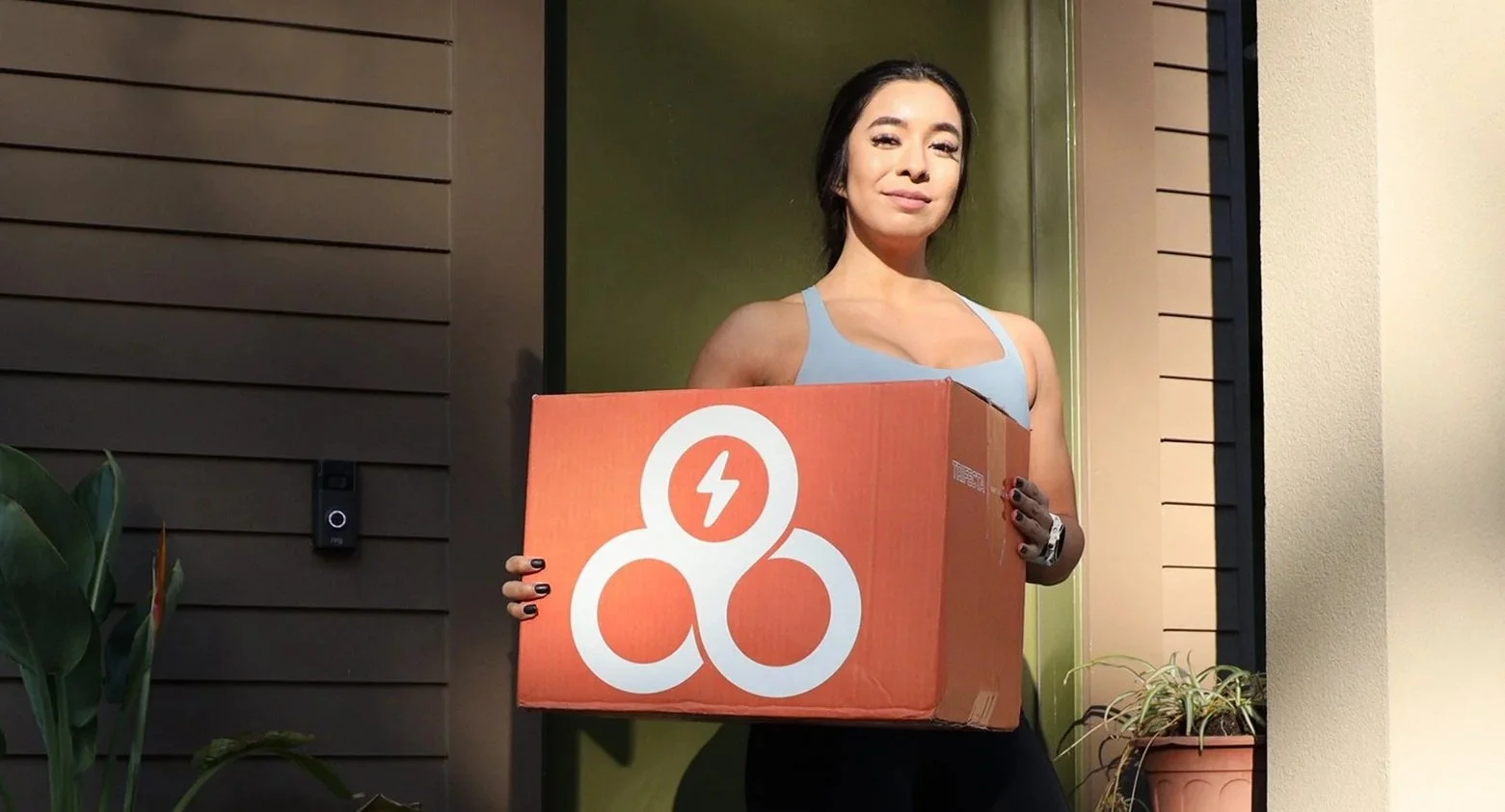 Woman standing outside holding a large cardboard box with a white logo featuring a lightning bolt inside a circle and three connected circles. She is smiling and wearing a light blue tank top, with plants and a house exterior visible in the background.