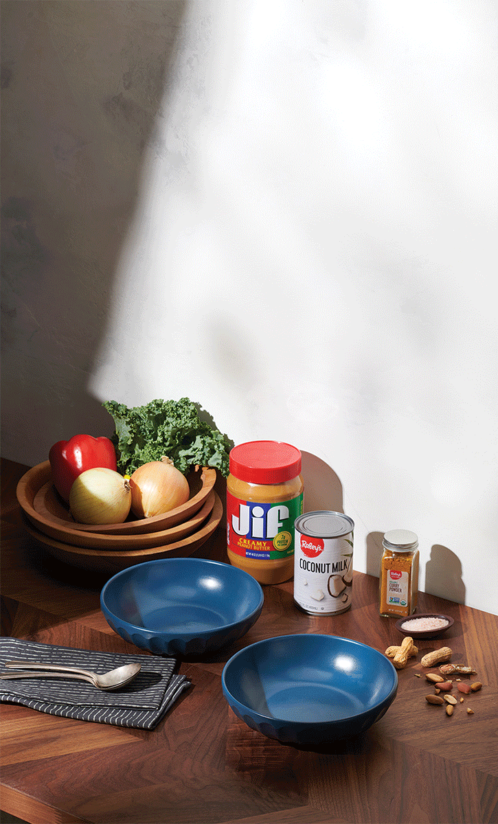 A wooden table with a blue bowl, silver spoon, black and white napkin, and various ingredients including red bell pepper, onions, kale, peanut butter, coconut milk, curry powder, and mixed nuts.
