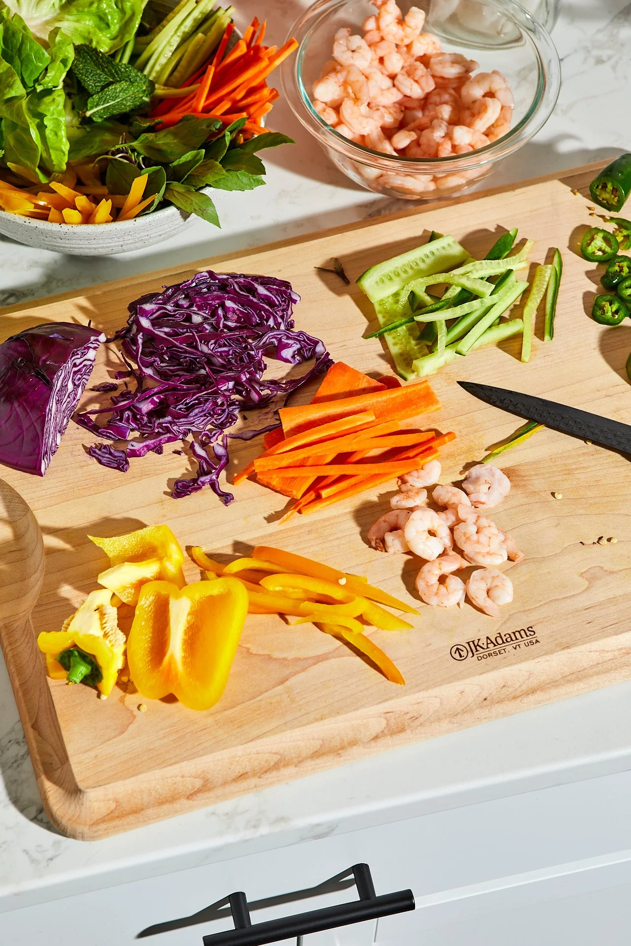 Chopped vegetables and shrimp on a wooden cutting board, including purple cabbage, cucumber, celery, jalapeño, yellow bell pepper, orange carrots, and cooked shrimp, with a bowl of additional shrimp and a bowl of assorted greens and vegetables in the