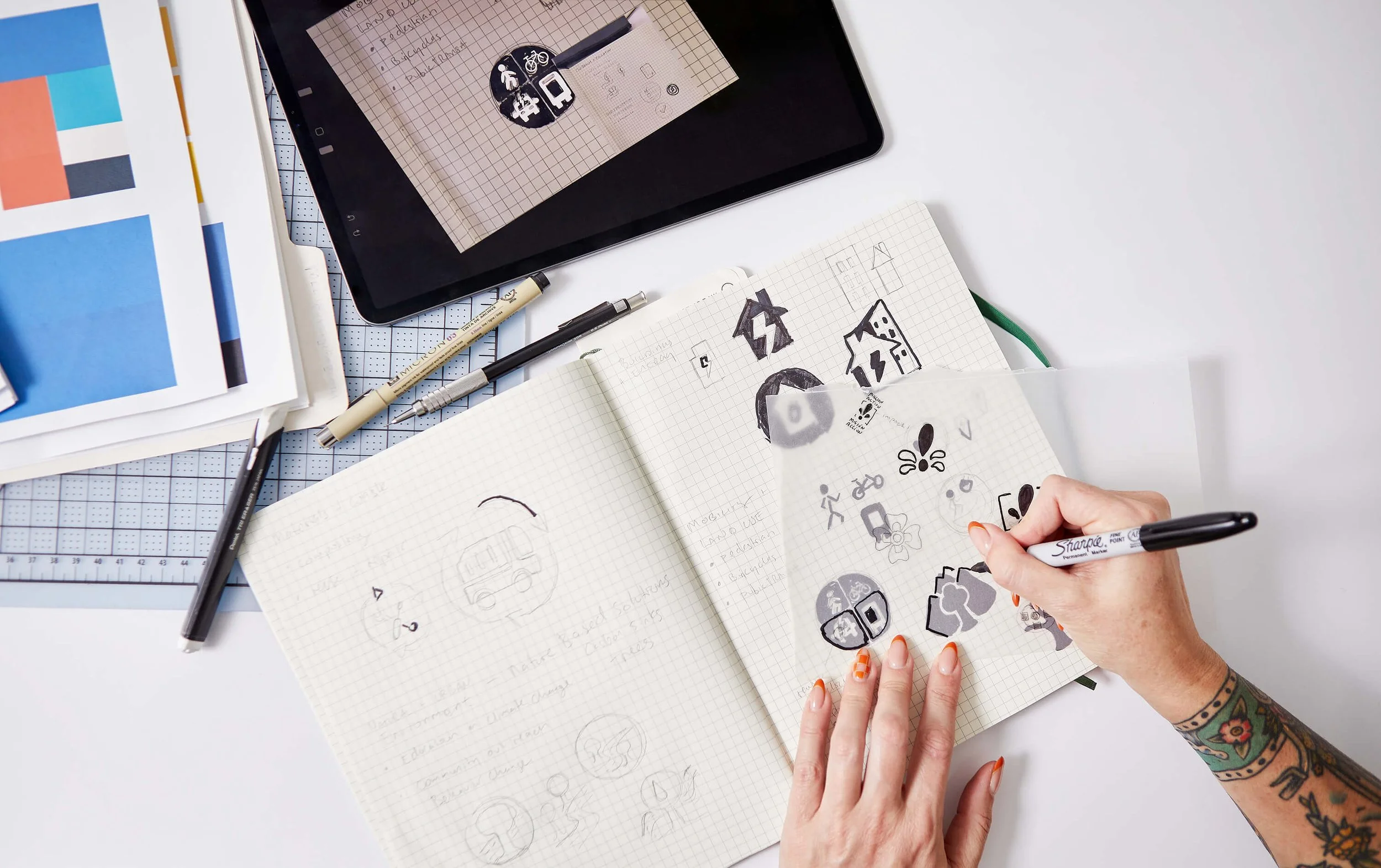 A workspace with notebooks, pens, a tablet, and printed color and grid sheets. A person with tattoos is sketching various icons and symbols on a grid notebook, surrounded by other drafting tools and visual references.