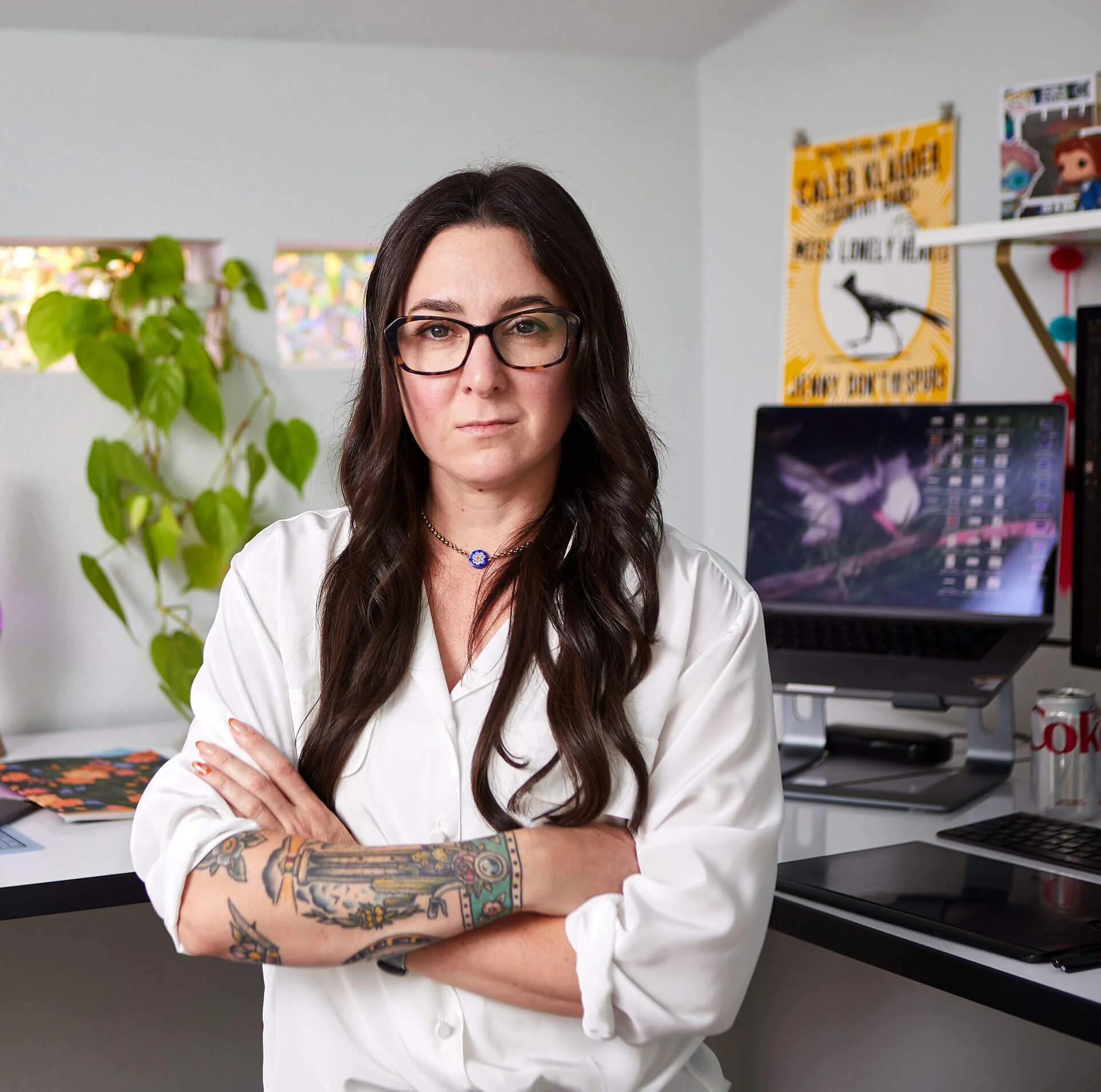 A woman with long dark hair, glasses, and tattoos on her arm stands with arms crossed in an office, wearing a white shirt. Behind her are computer monitors, a soda can, and a yellow poster with a bird and text.
