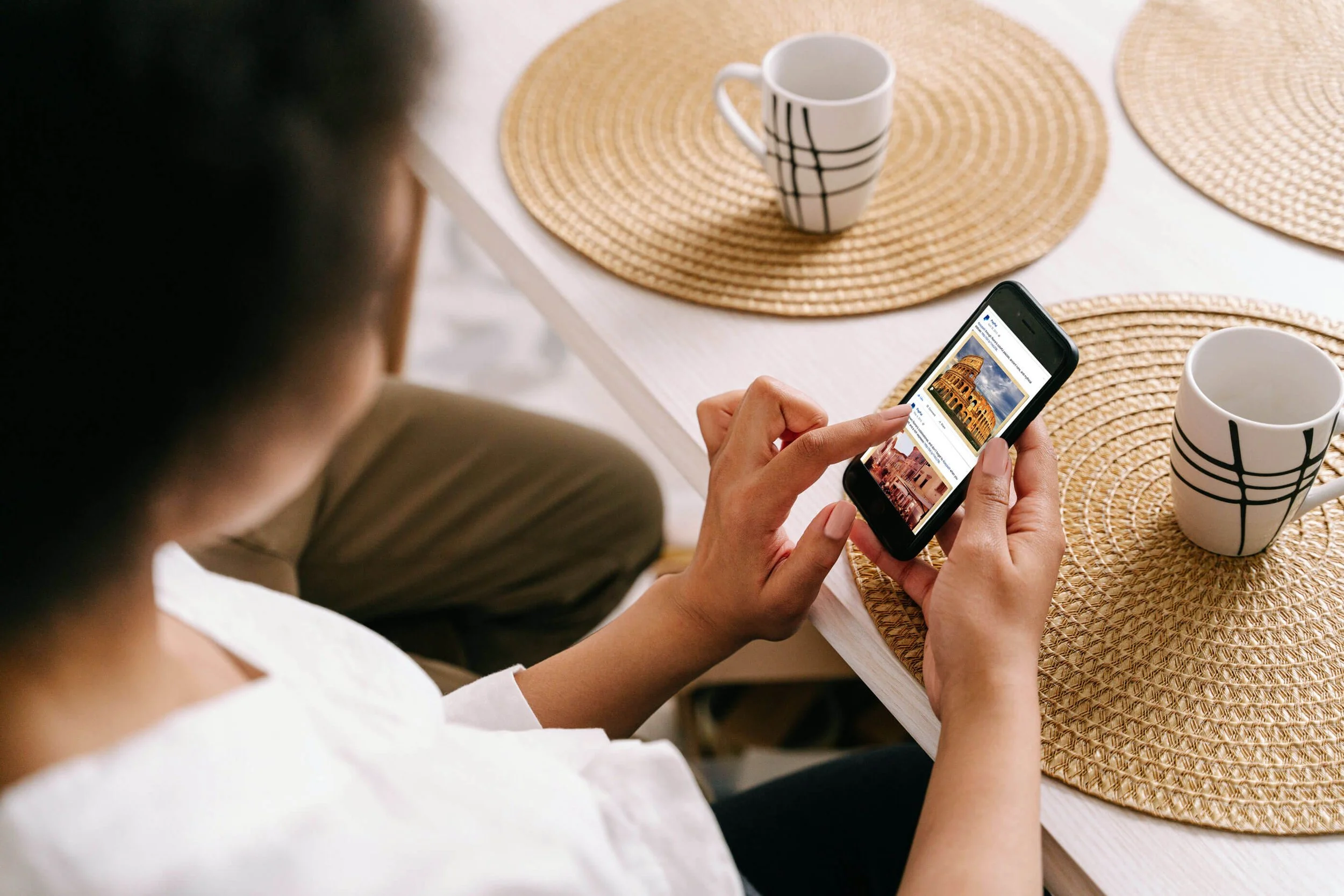 Woman sitting at a table using a smartphone, with two coffee mugs and woven placemats on the table.
