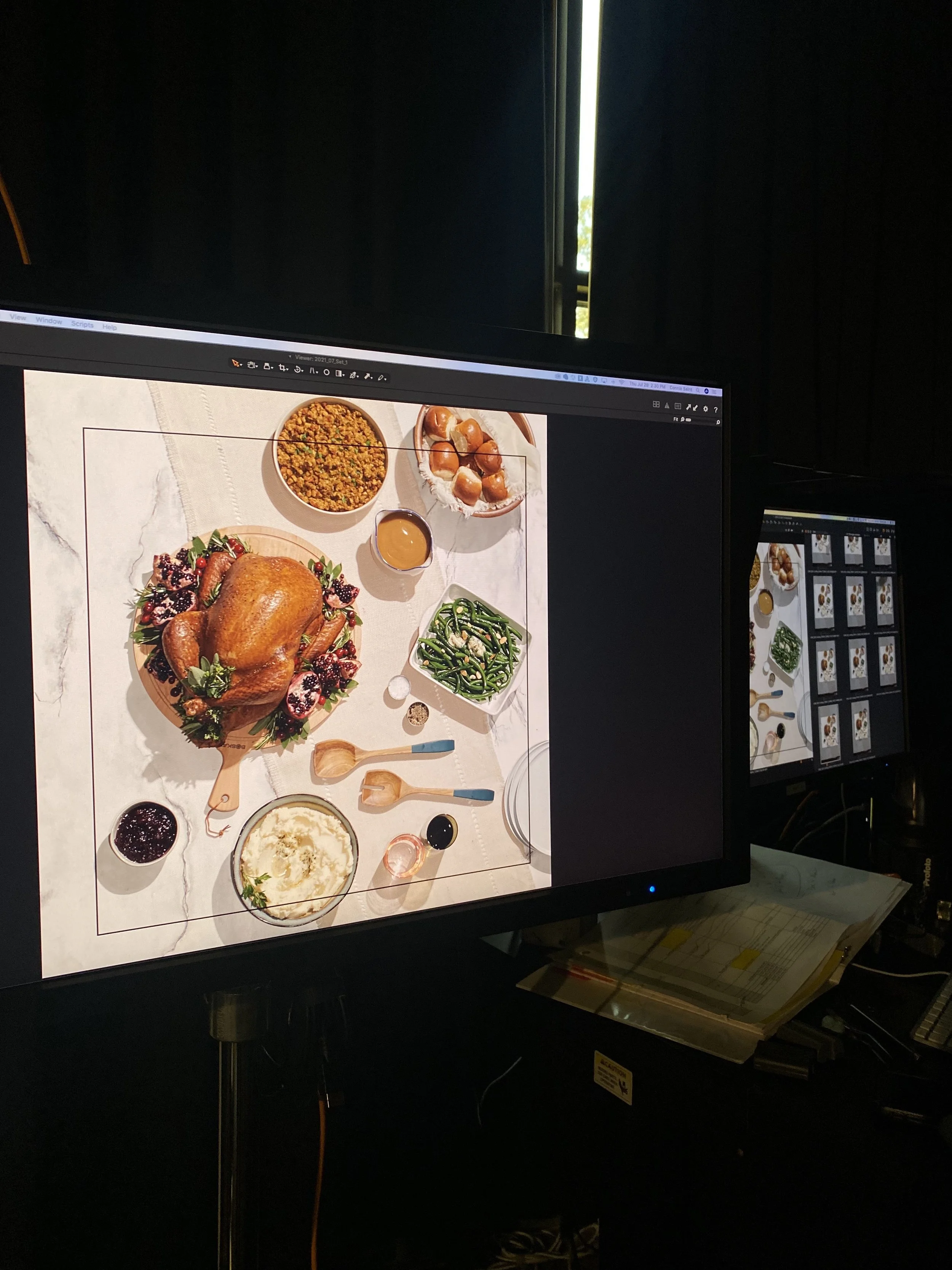 Computer monitor displaying a photo of a Thanksgiving feast with a roasted turkey, stuffing, green beans, mashed potatoes, cranberry sauce, gravy, bread rolls, and various condiments on a white tablecloth.