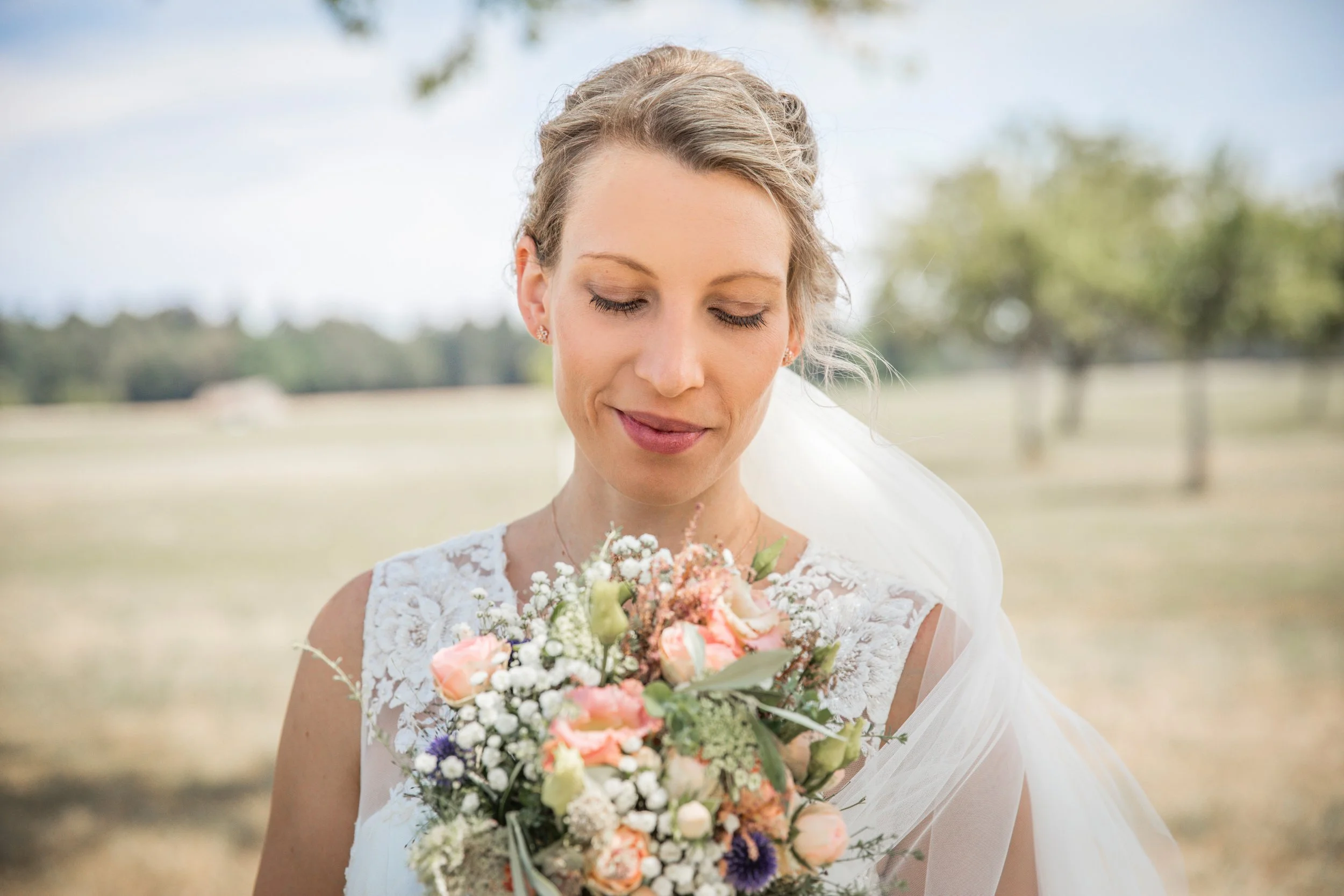 Braut hält einen Blumenstrauß, steht im Freien, trägt ein weißes Hochzeitskleid, hat blonde, lockige Haare und schaut auf den Blumenstrauß.