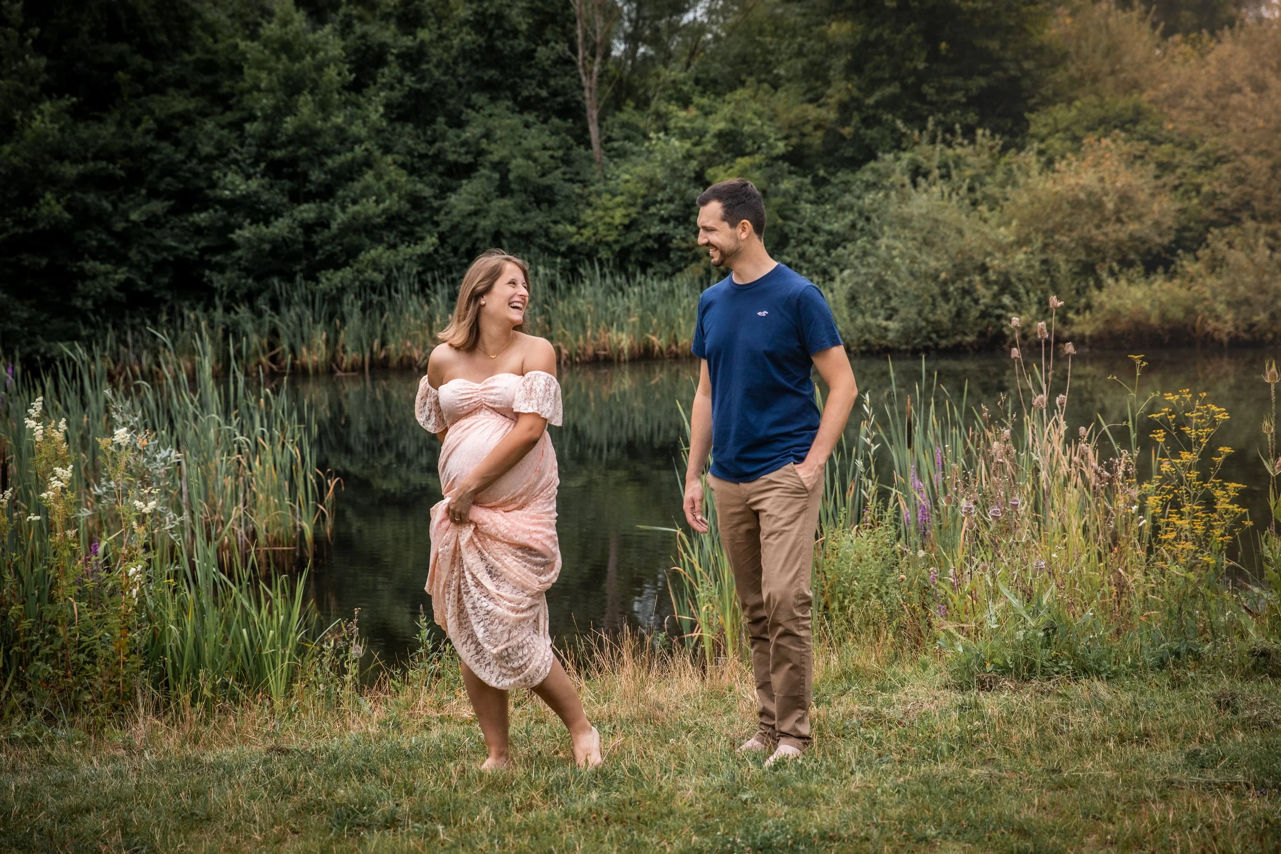 Ein lachendes Paar, eine schwangere Frau in einem rosa Kleid, steht an einem See im Grünen, bei Sonnenuntergang, im Freien.