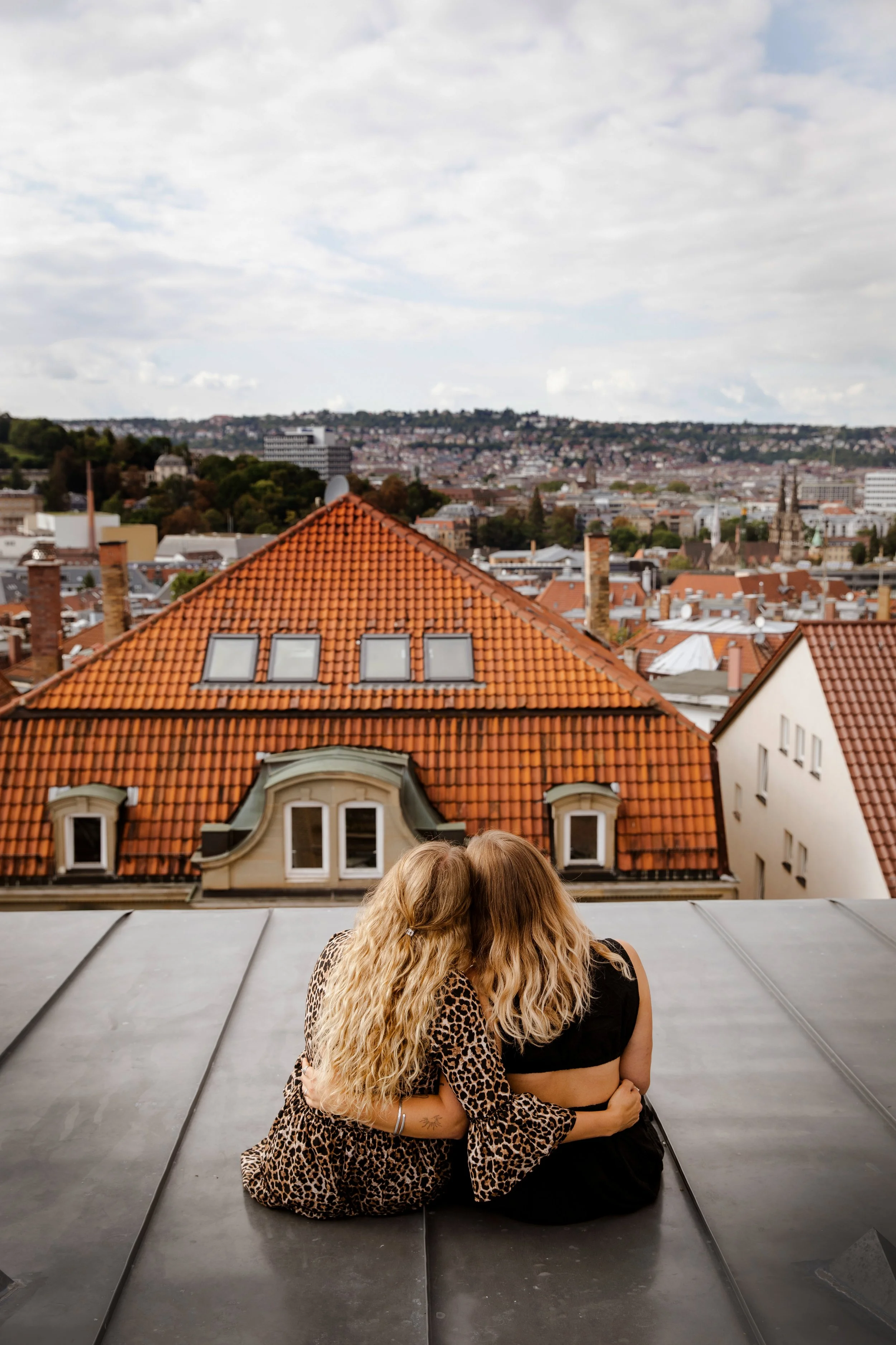 Zwei Frauen sitzen auf einem Flachdach mit Blick auf eine Stadt mit roten Dächern und Hügeln im Hintergrund, während sie sich umarmen.