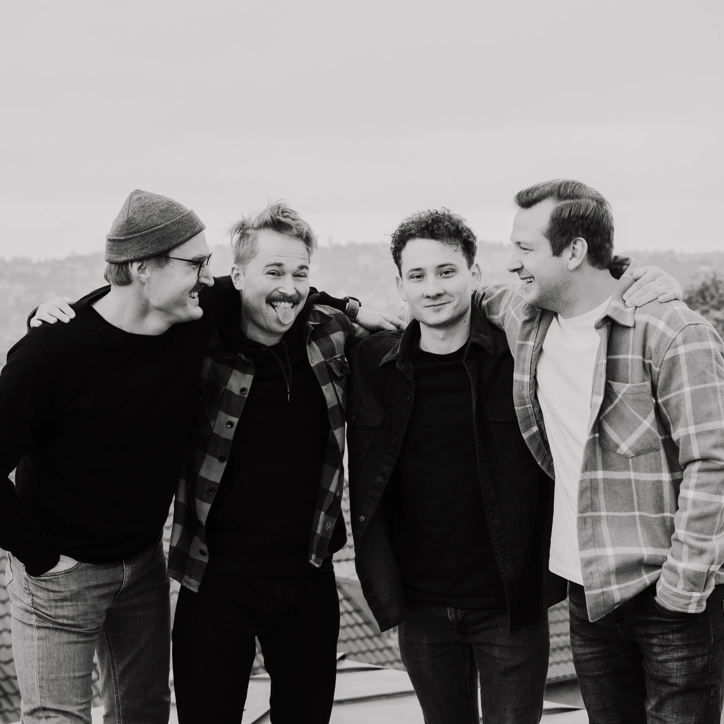 Fünf junge Männer auf einem Dach, die zusammen lachen und sich umarmen, bei Sonnenuntergang, Schwarz-Weiß-Foto.