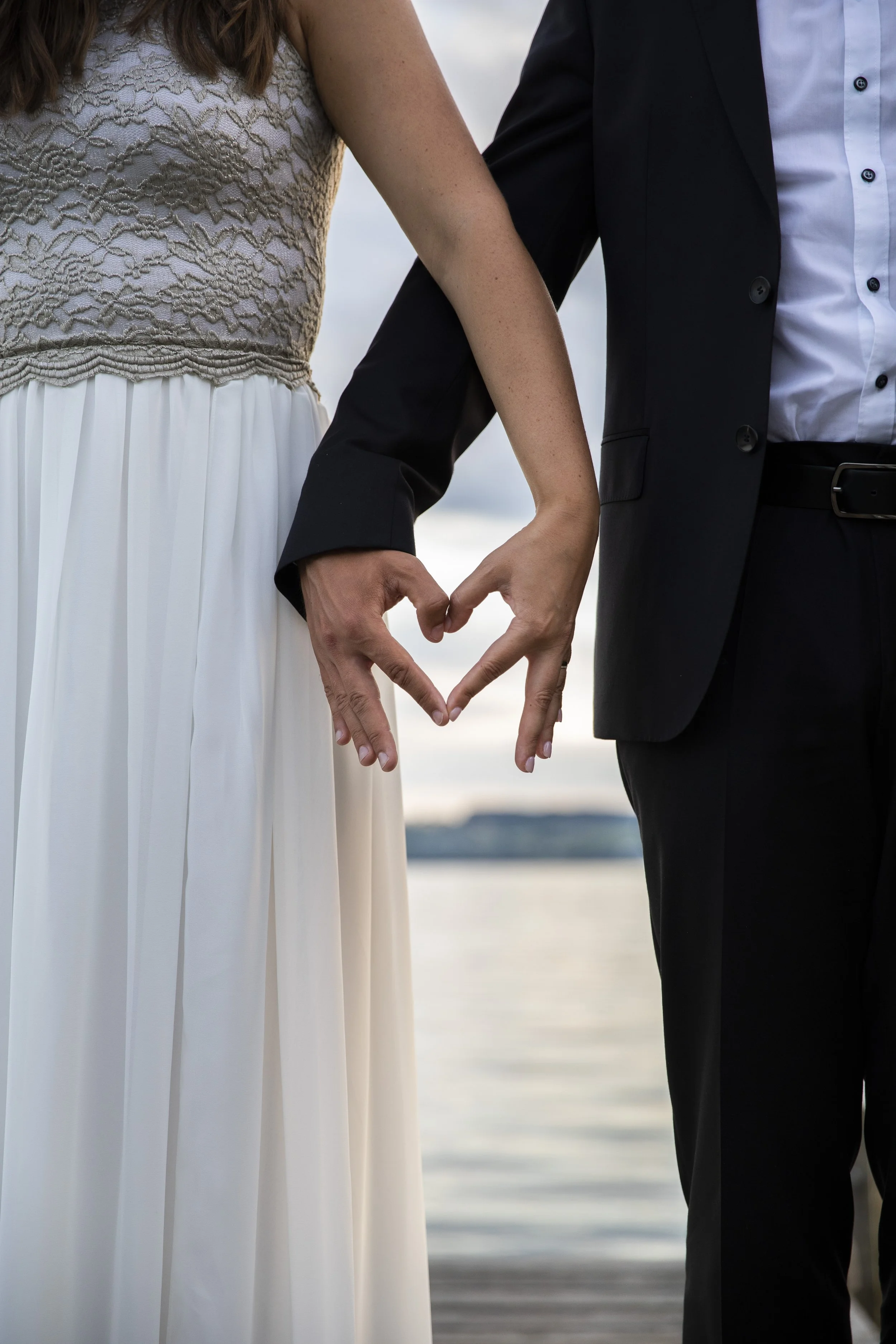 Ein Paar hält sich an den Händen, um eine Herzform zu bilden, während sie am Wasser stehen, vermutlich an ihrem Hochzeitstag.