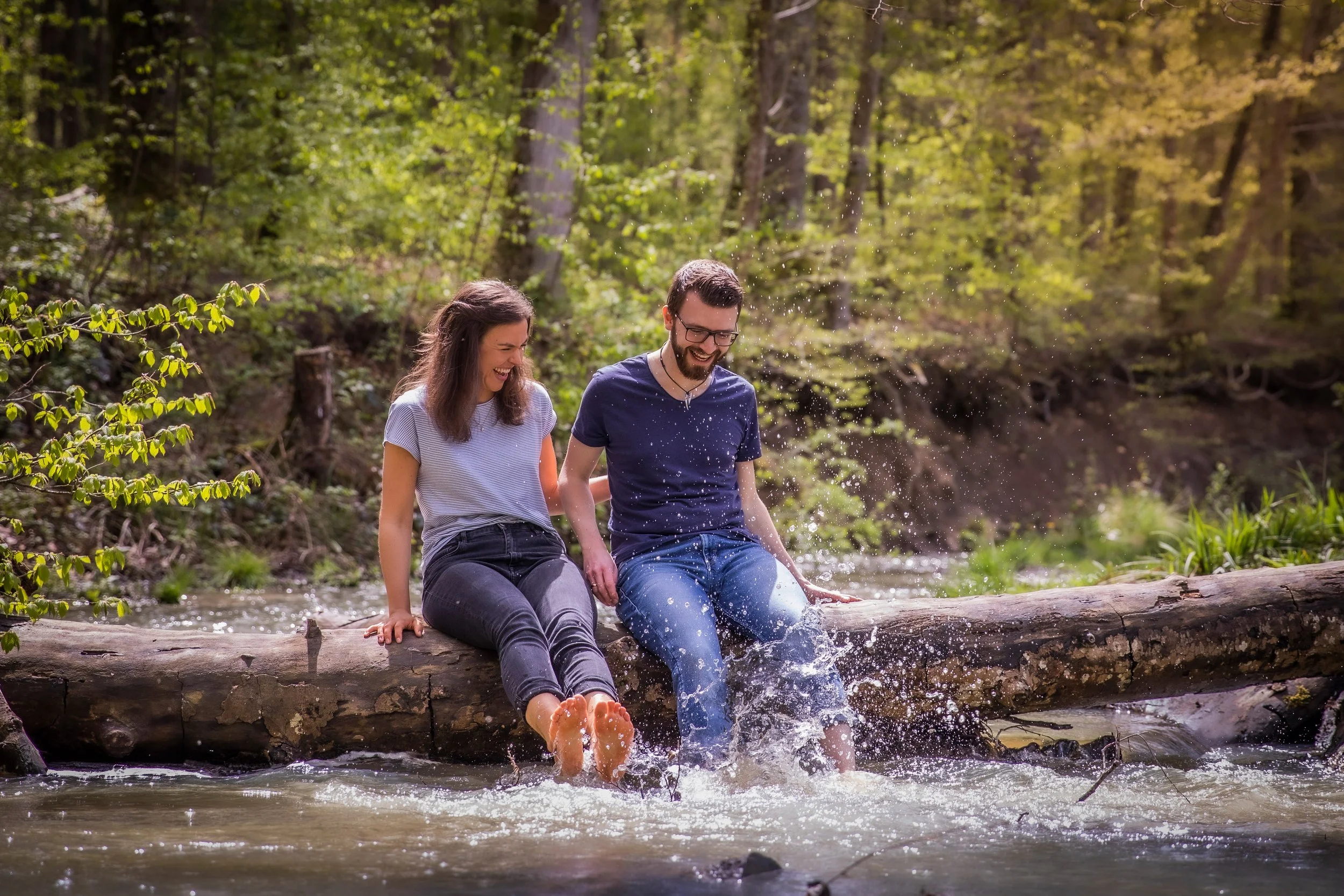 Ein Mann und eine Frau sitzen auf einem Baumstamm im Wasser eines Flusses im Wald und spielen mit dem Wasser, lachen und haben Spaß.
