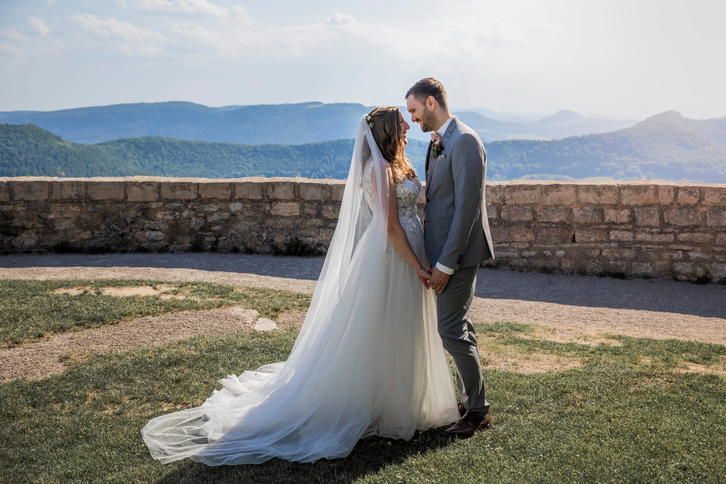 Ein Brautpaar in Hochzeitskleidung hält Händchen auf einer Wiese mit Berge im Hintergrund.