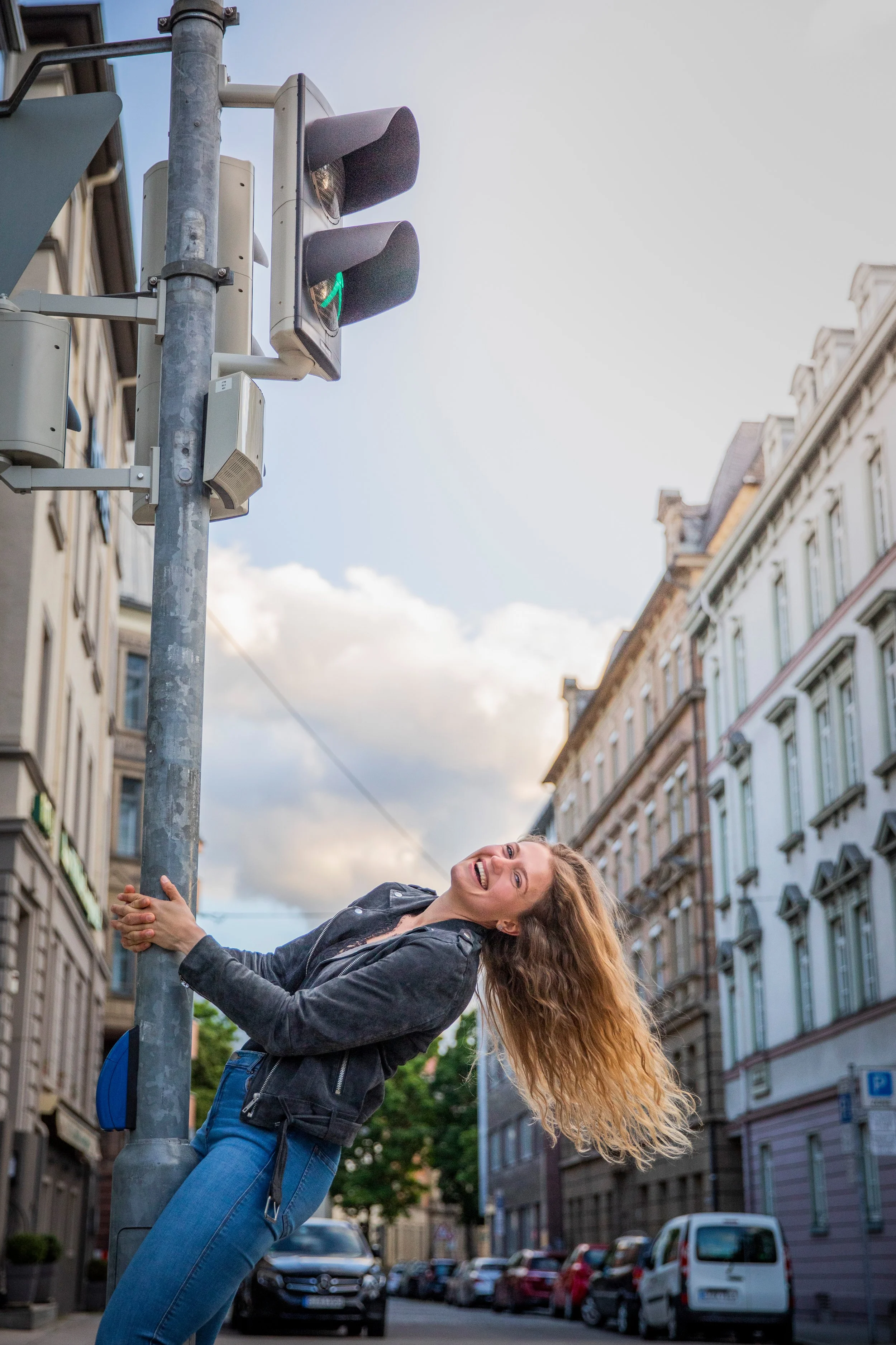 Eine lachende junge Frau in dunkler Jacke und blauen Jeans umarmt einen Poller an einer Stadtstraße mit alten Gebäuden und parkierten Autos, während sie den Blick gegen die Kamera richtet.