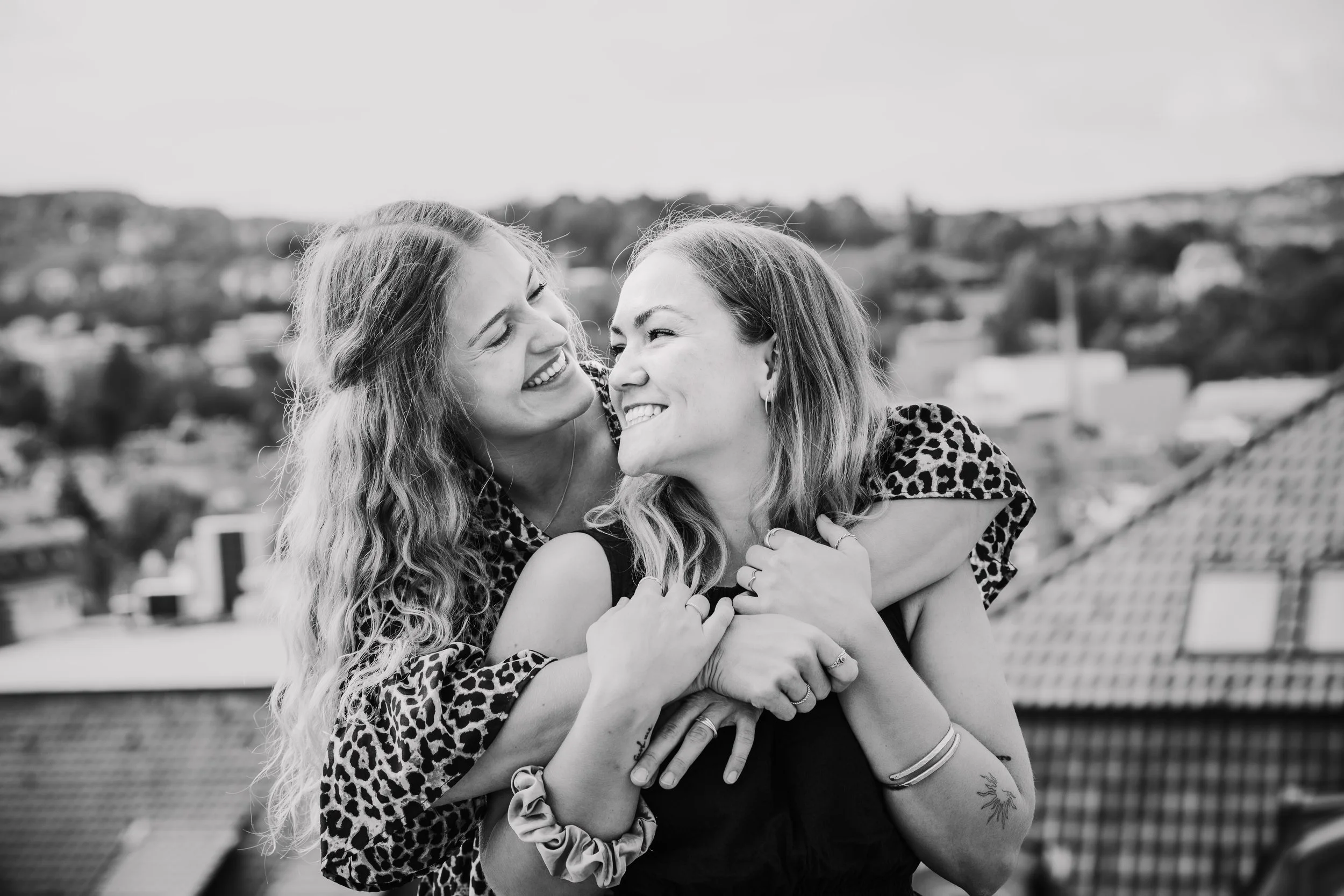 Zwei Frauen umarmen sich lachend auf einem Balkon, im Hintergrund eine Stadtlandschaft, schwarz-weiß Foto.