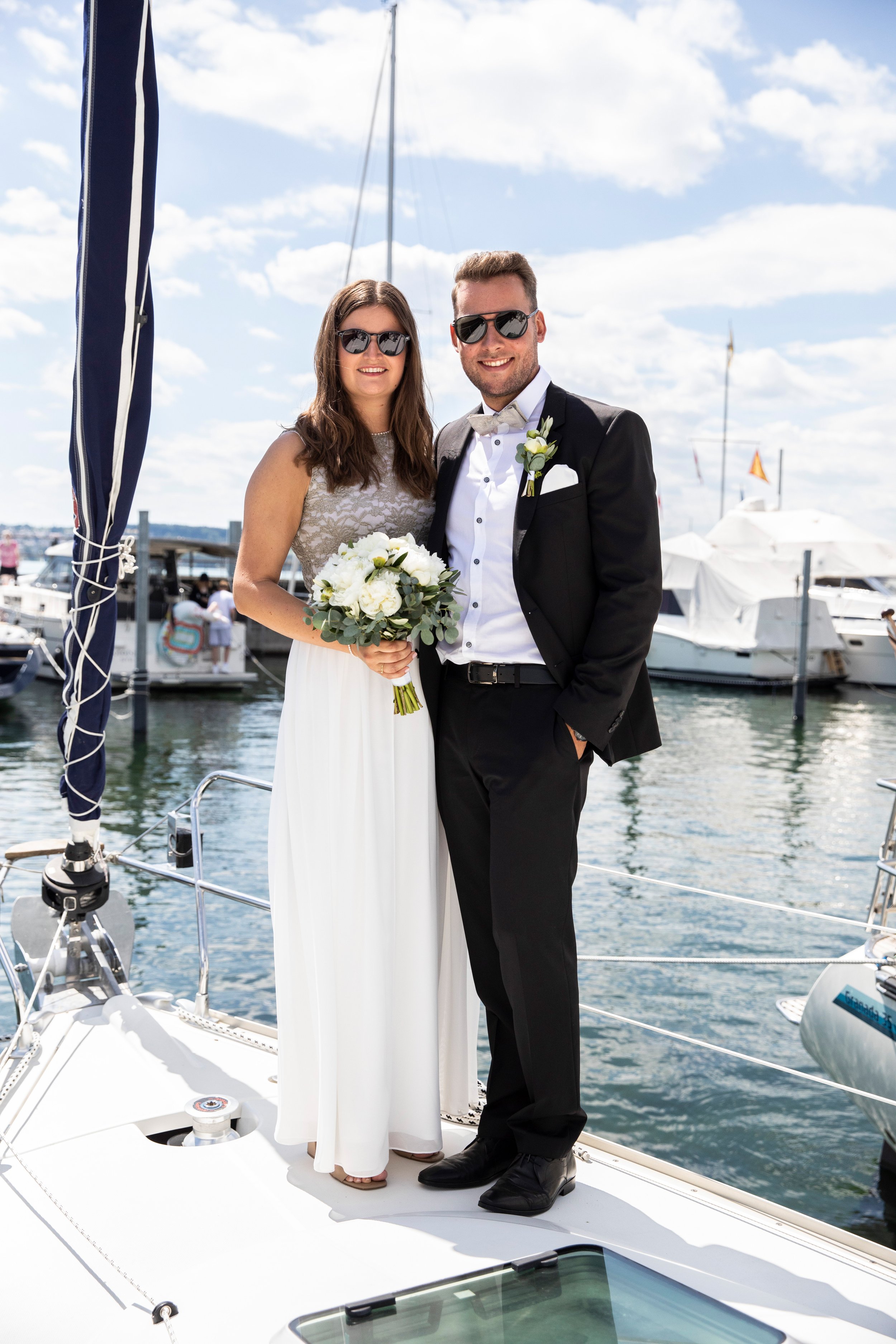Ein verheiratetes Paar in formeller Kleidung auf einem Boot am Wasser, mit Segel und anderen Booten im Hintergrund, bei gutem Wetter mit Wolken am Himmel.