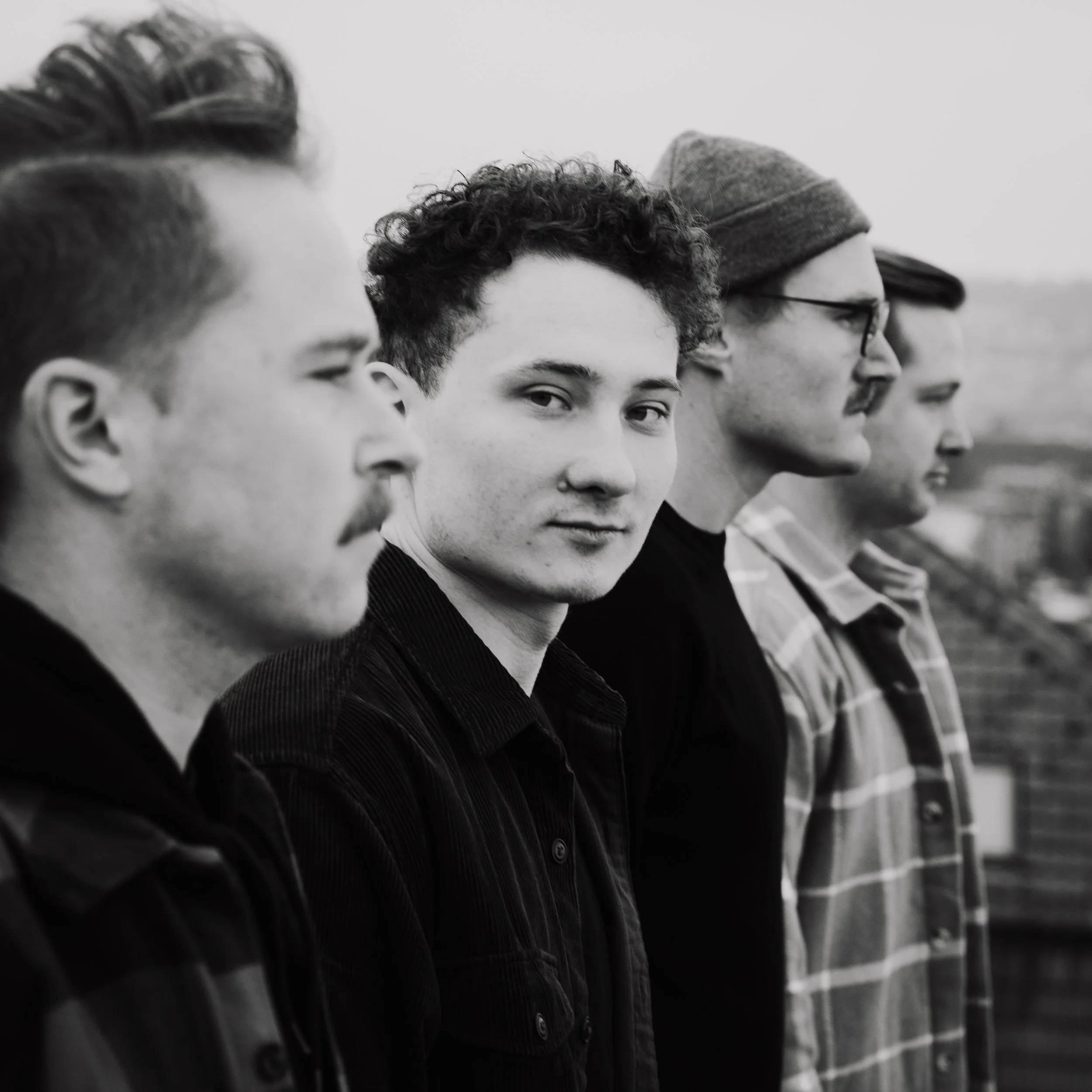 Fünf junge Männer stehen in einer Reihe, schauen in unterschiedliche Richtungen, auf einem Feld bei bewölktem Himmel, in Schwarz-Weiß fotografiert.
