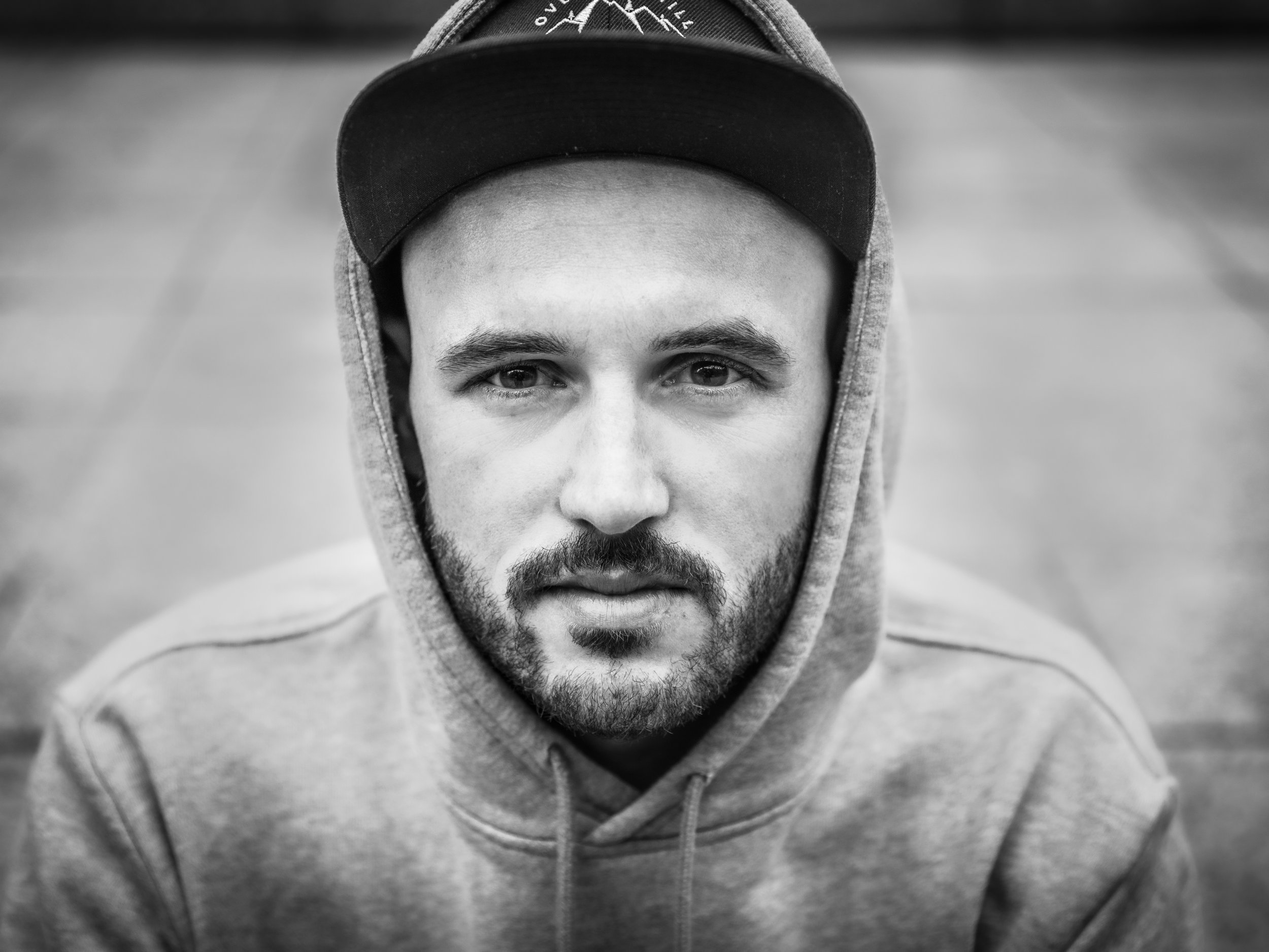 Schwarz-weiß Porträt eines jungen Mannes mit Hoodie und Cap, direkte Blick in die Kamera, unscharfer Hintergrund.