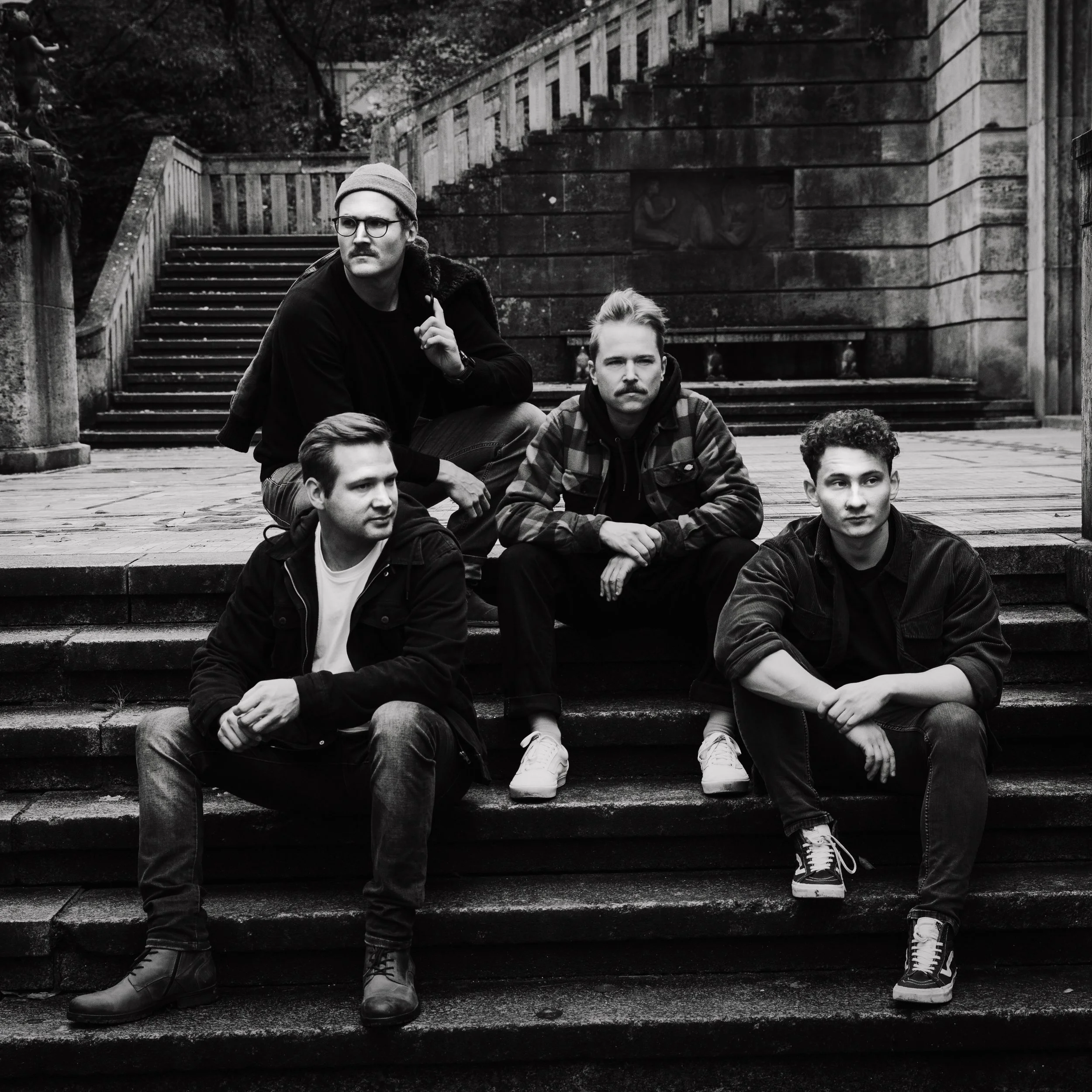 Fünf junge Männer sitzen auf einer Treppe im Freien, schwarz-weiße Fotografie, moderne Kleidung, urbanes Umfeld, seriöser Ausdruck, Gruppe, entspannte Atmosphäre.