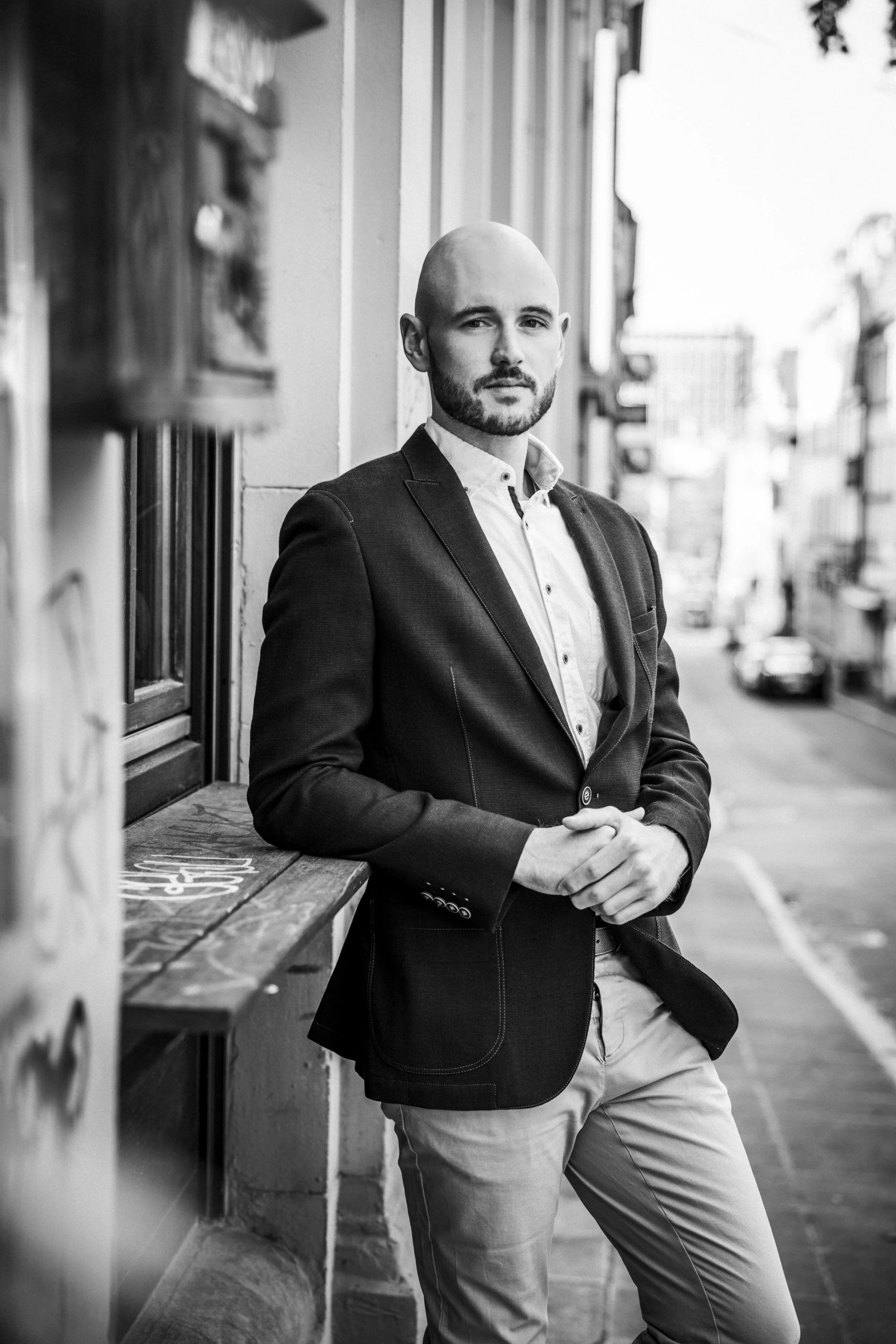 Schwarz-weiß-Foto eines Mannes in einem Anzug, der an einer Hauswand in einer Stadtstraße lehnt.