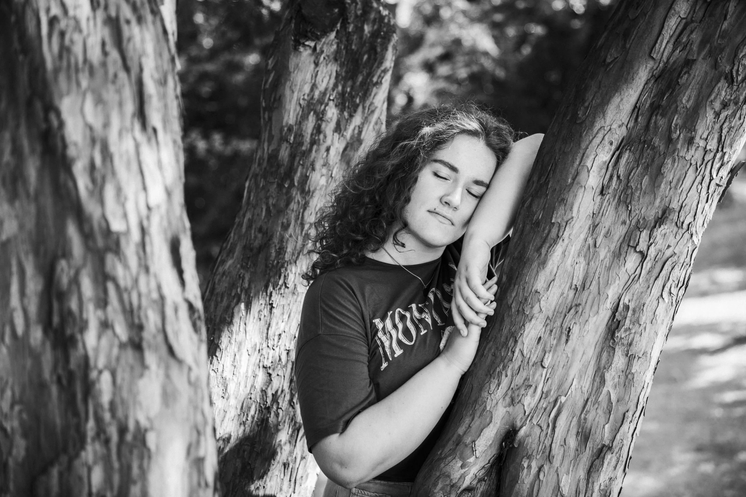 Junge Frau mit lockigem Haar lehnt an einem Baum, die Augen geschlossen, in einer natürlichen Umgebung, Schwarz-Weiß-Fotografie.