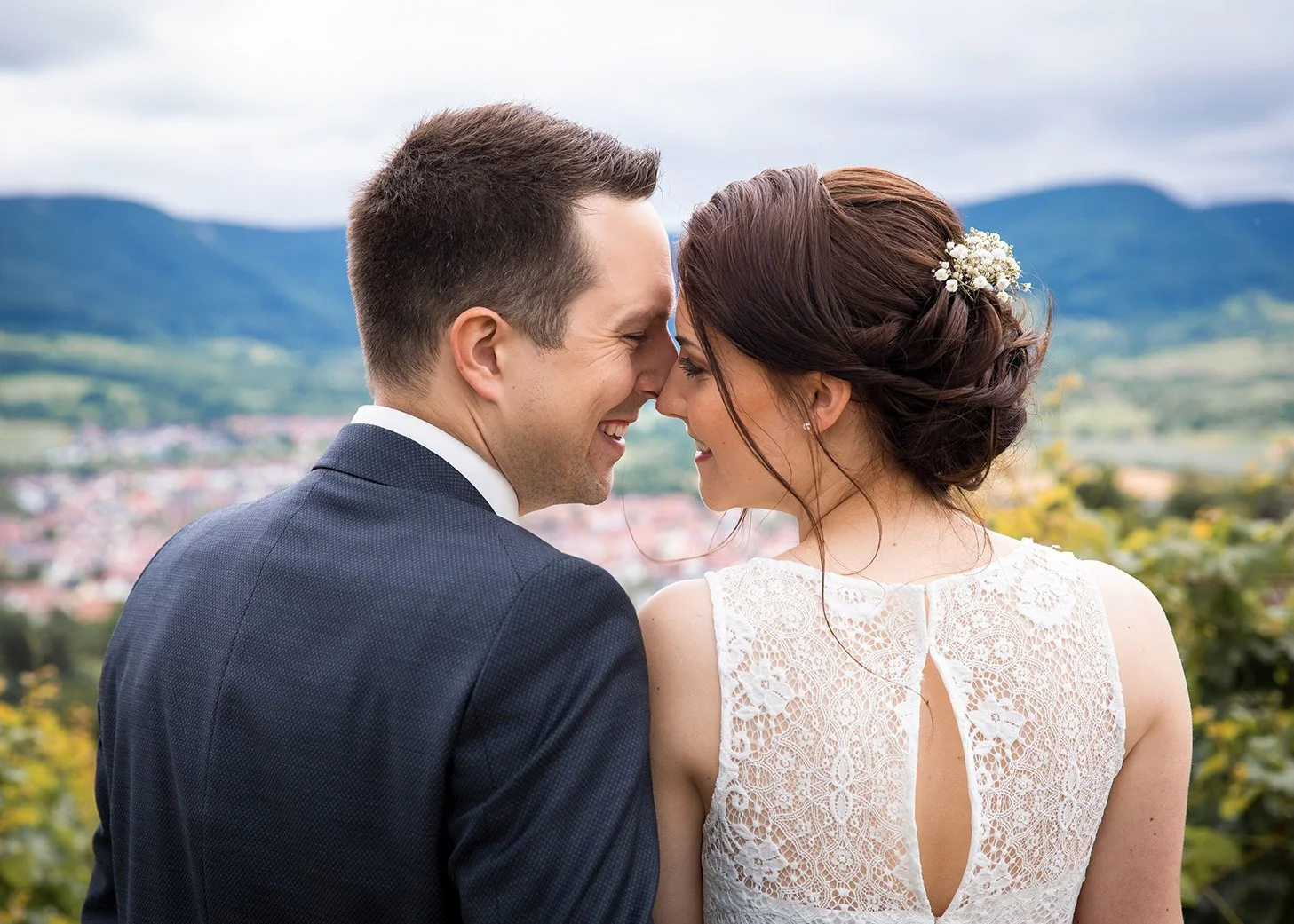 Ein glückliches Brautpaar steht eine Seite nebeneinander, lächelnd und ihre Stirn berührend, mit einem malerischen Berg- und Landschaftshintergrund.
