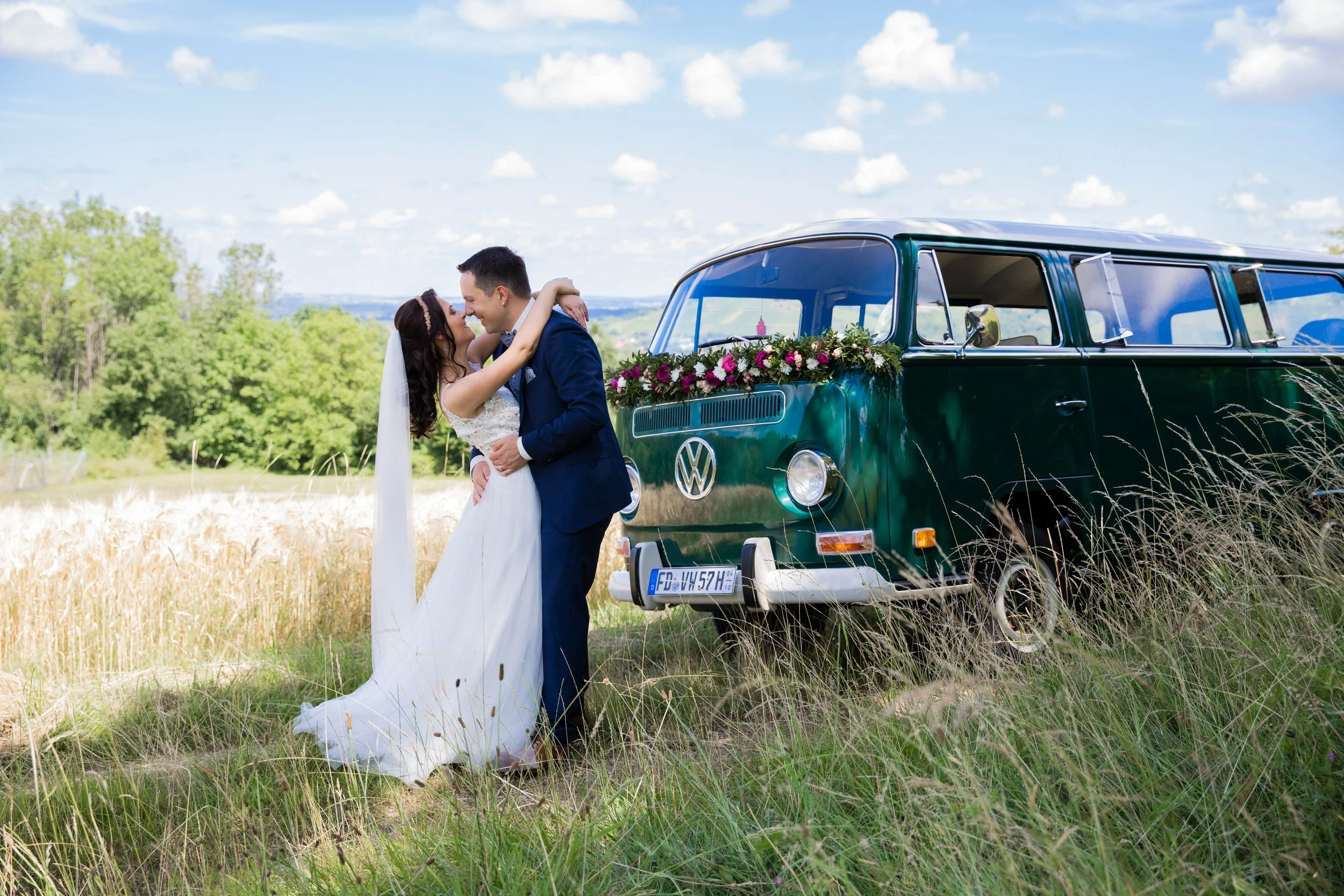 Ein frisch verheiratetes Paar freut sich in einem Feld neben einem grünen VW-Bropan, das mit Blumen dekoriert ist, bei schönem Wetter