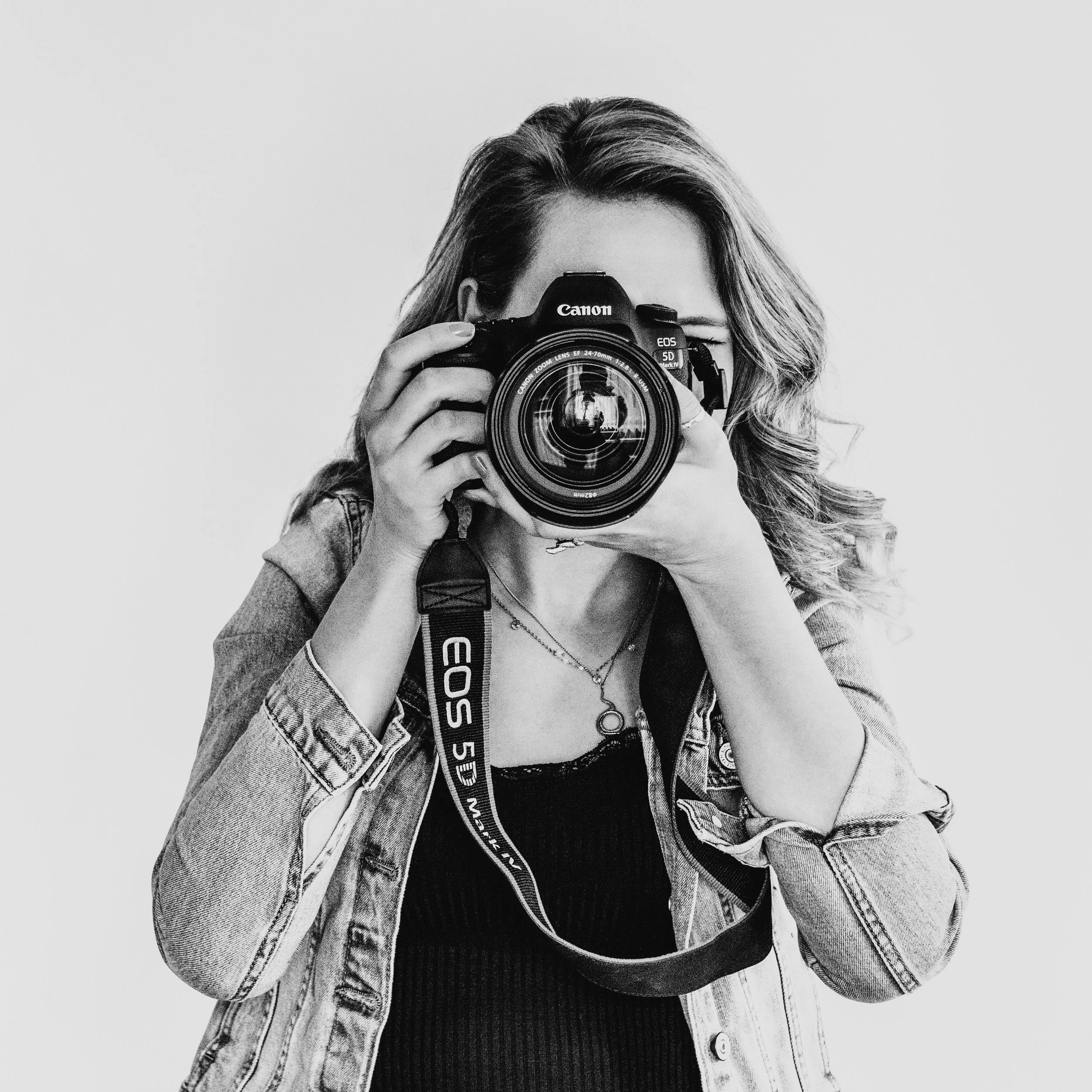 Schwarz-Weiß-Foto einer jungen Frau, die eine Kamera vor das Gesicht hält, mit lockigem Haar und einem Jeansjacke