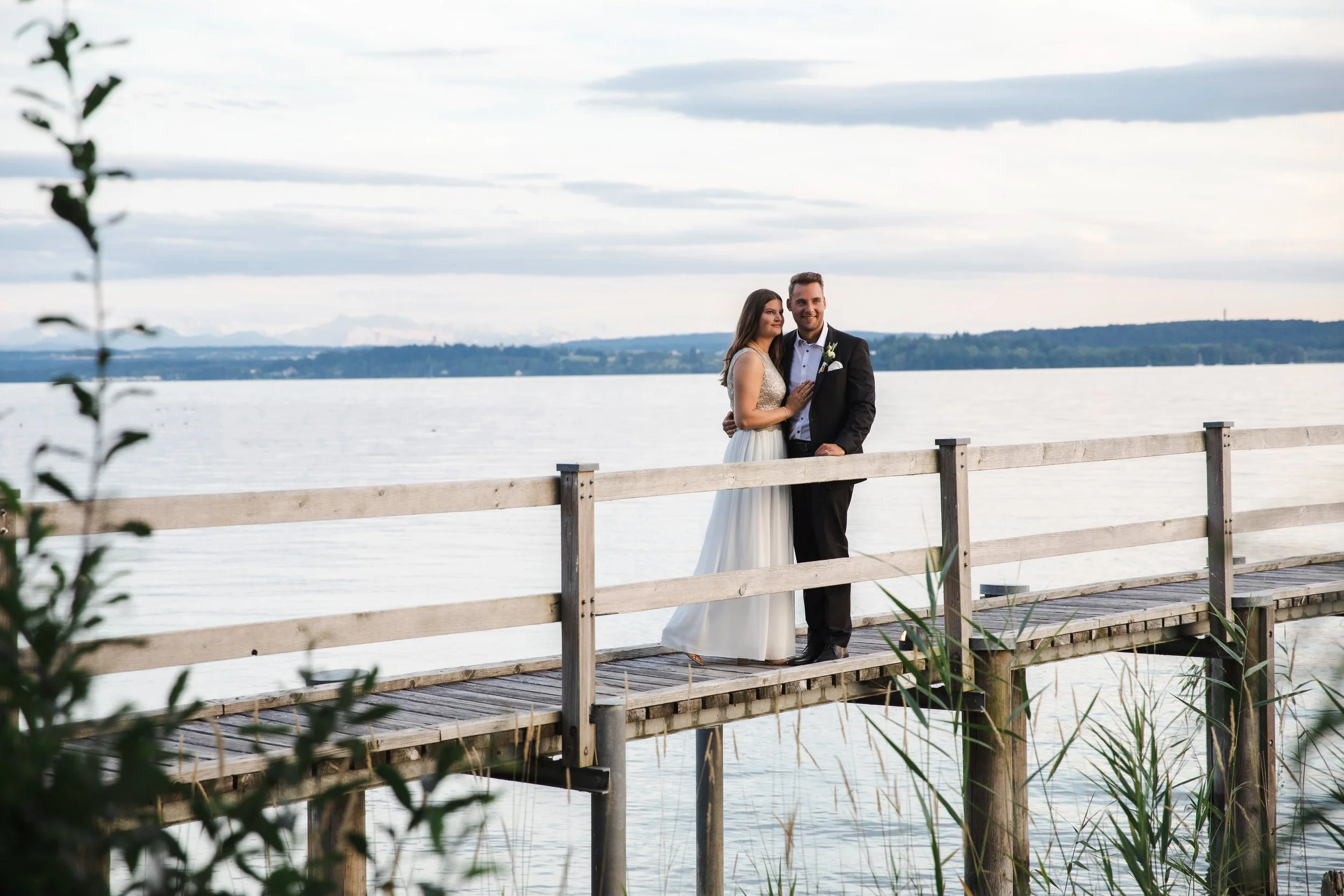 Ein frisch vermähltes Paar steht auf einer wooden Plattform am See, im Hintergrund Wasser und Landschaft, bei bewölktem Himmel.