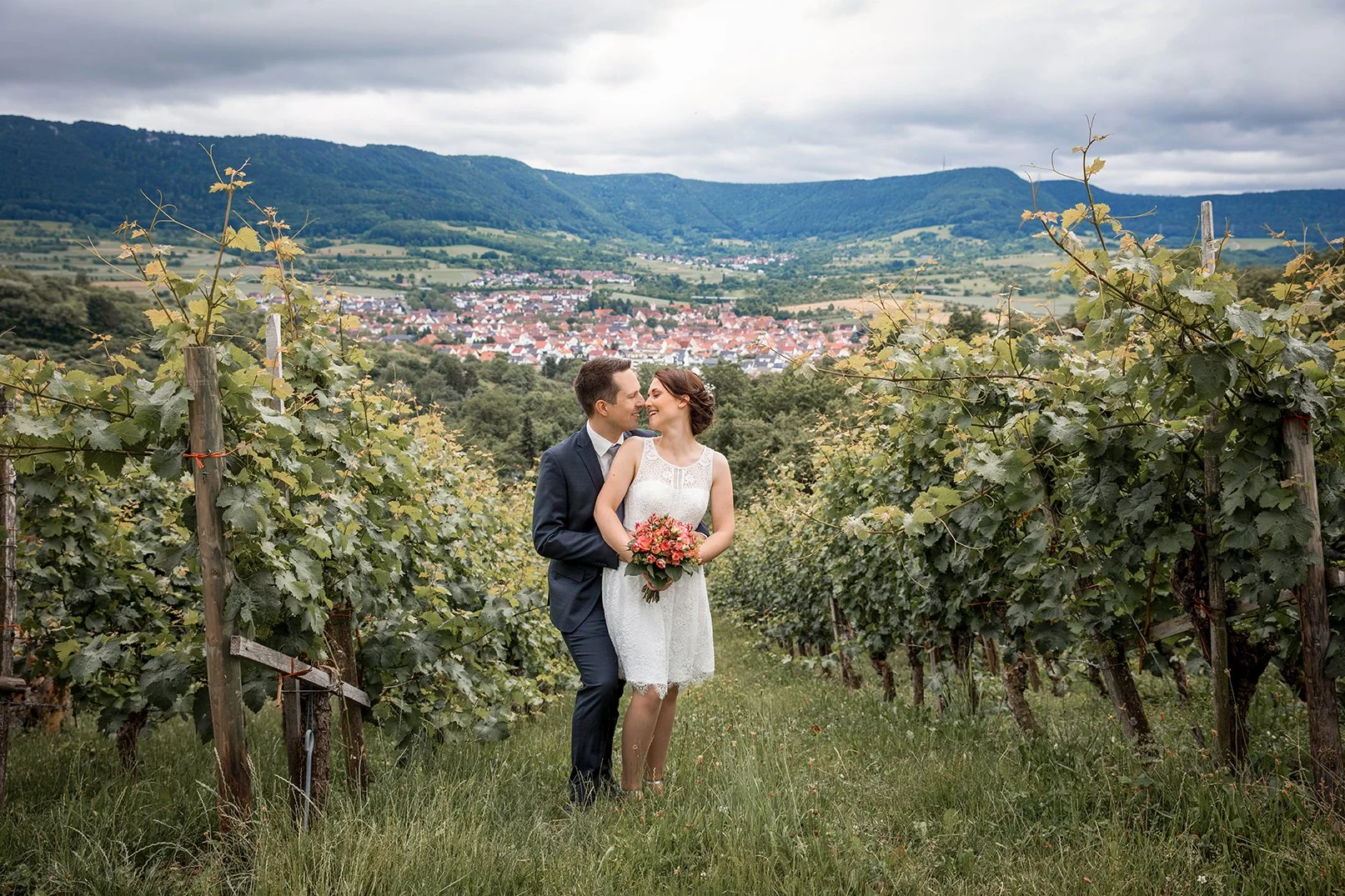 Ein frisch verheiratetes Paar in einem Weinberg mit Blick auf ein Dorf in den Hügeln, umgeben von Weinreben, an einem bewölkten Tag.