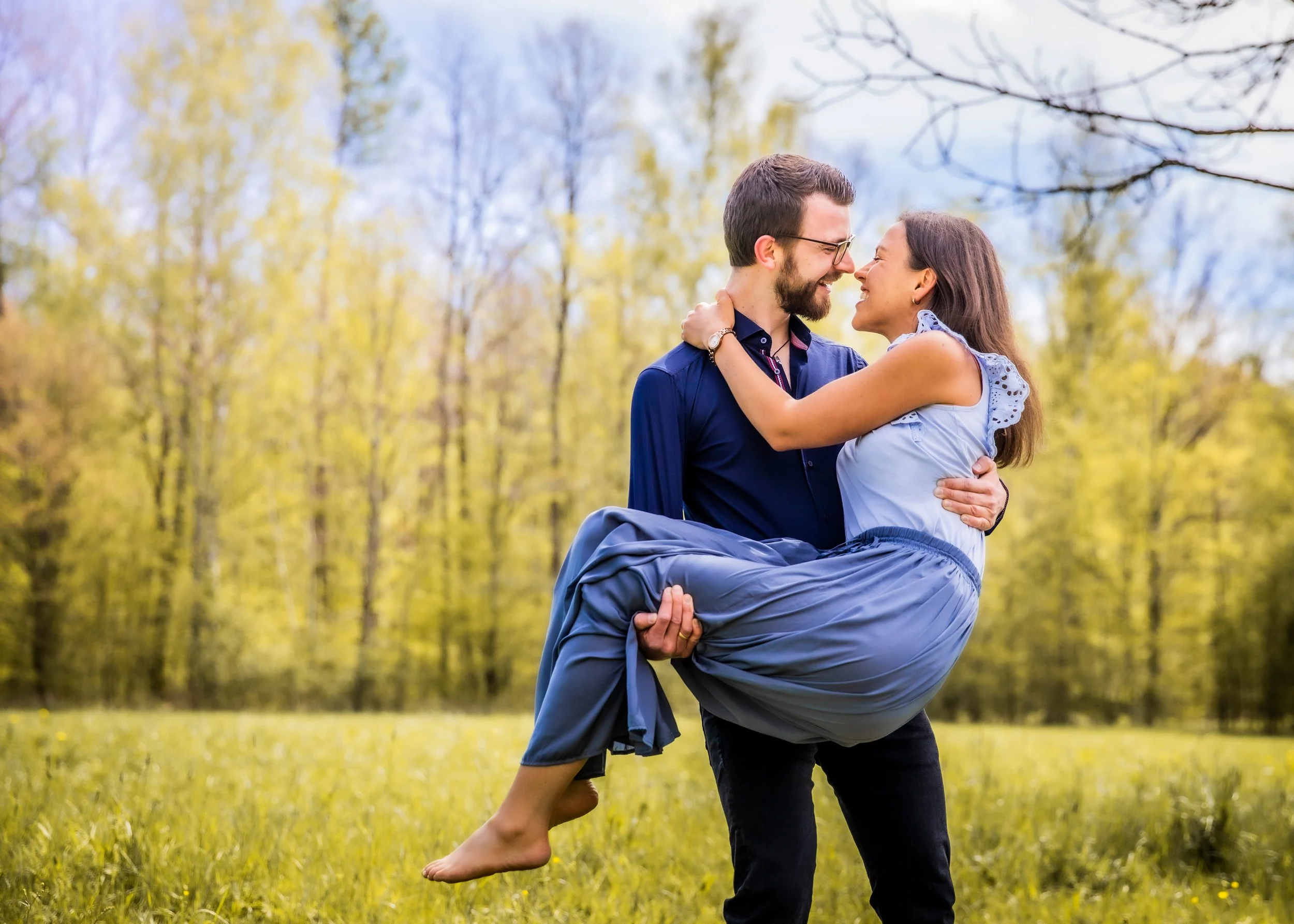 Ein lachendes Paar im Park, der Mann trägt eine dunkle Jacke, die Frau trägt eine hellblaue Bluse und einen hellblauen Rock, der Mann trägt schwarze Hosen, die Frau sitzt auf den Armen des Mannes, im Hintergrund Bäume mit frischem grünem Laub.