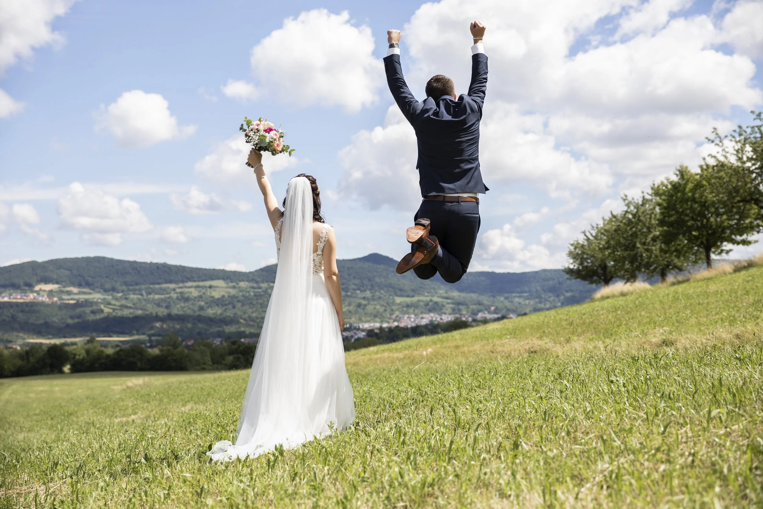 Hochzeitspaar im Freien, der Bräutigam springt in die Luft, die Braut hält einen Blumenstrauß, die Landschaft ist grün mit Hügeln und Bäumen, ein blauer Himmel mit Wolken.