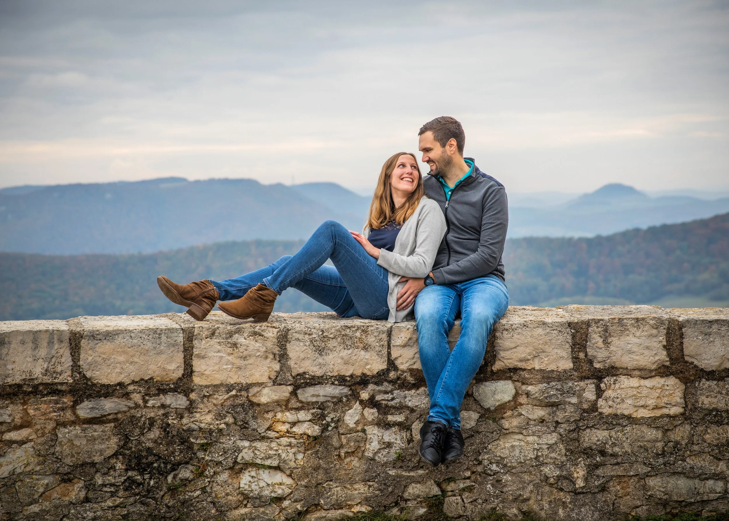 Ein Paar sitzt auf einer Steinmauer vor einer bergigen Landschaft, lächelt und schaut sich verliebt an.